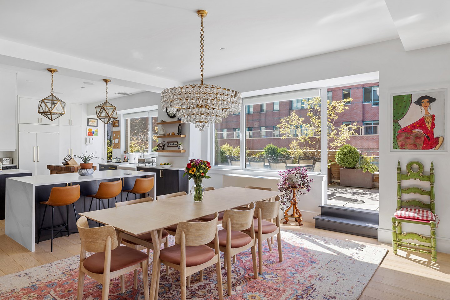 Bright, modern kitchen and dining area with large windows, a wooden dining table with chairs, a chandelier, and a kitchen island with stools. Decor includes colorful paintings, potted plants, and a patterned rug.