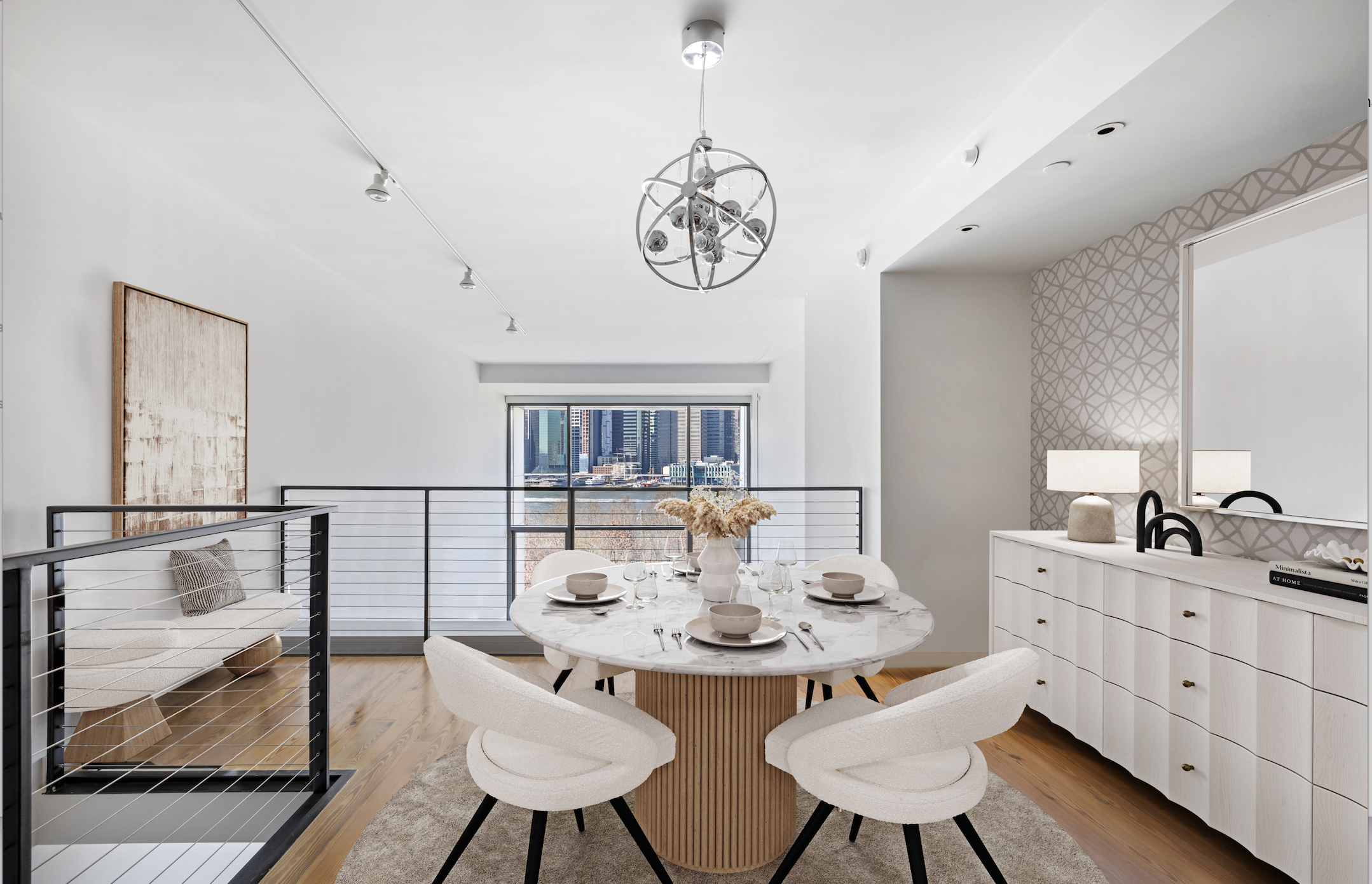 Modern dining room with a marble oval table set with four bowls, glasses, and cutlery, surrounded by four cream-colored chairs, a white sideboard with lamps and decor, large window showing city buildings, and a pendant light fixture hanging from the ceiling.