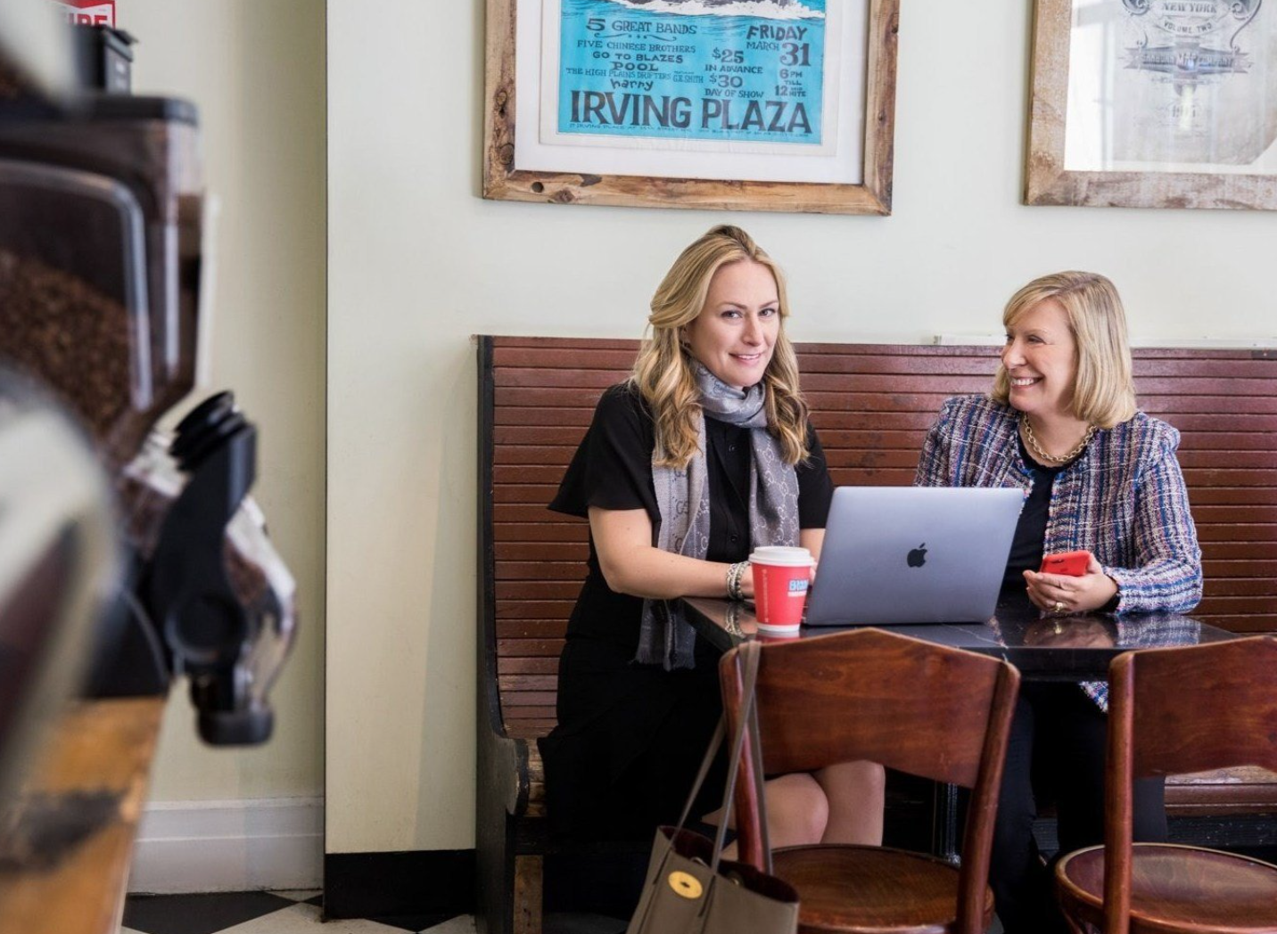 Two women sitting at a coffee shop table, smiling and talking. One has a MacBook laptop open in front of her, and a red coffee cup on the table. The other is holding a red phone, wearing a colorful blazer. The background has framed posters on a white