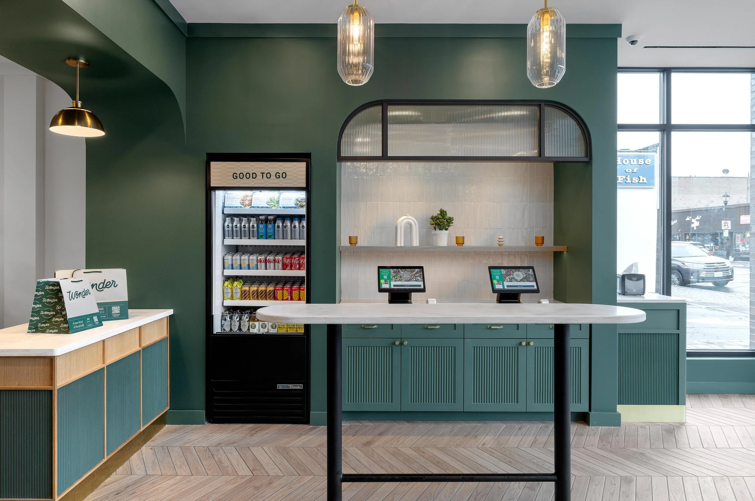 Interior of a modern coffee shop with green walls, a white countertop, digital ordering screens, and a beverage refrigerator. Large windows show a street outside.