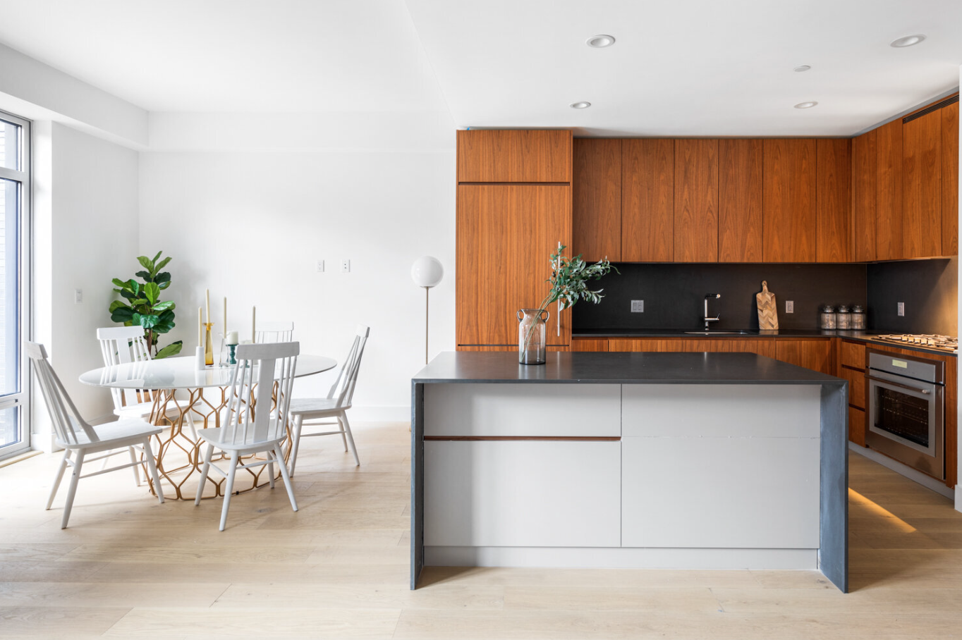 Modern kitchen and dining area with white walls, light wood flooring, a round dining table with white chairs, a potted plant, and a kitchen island with a black countertop and a vase with greenery. The kitchen has wooden cabinets, a black backsplash, and stainless steel appliances.