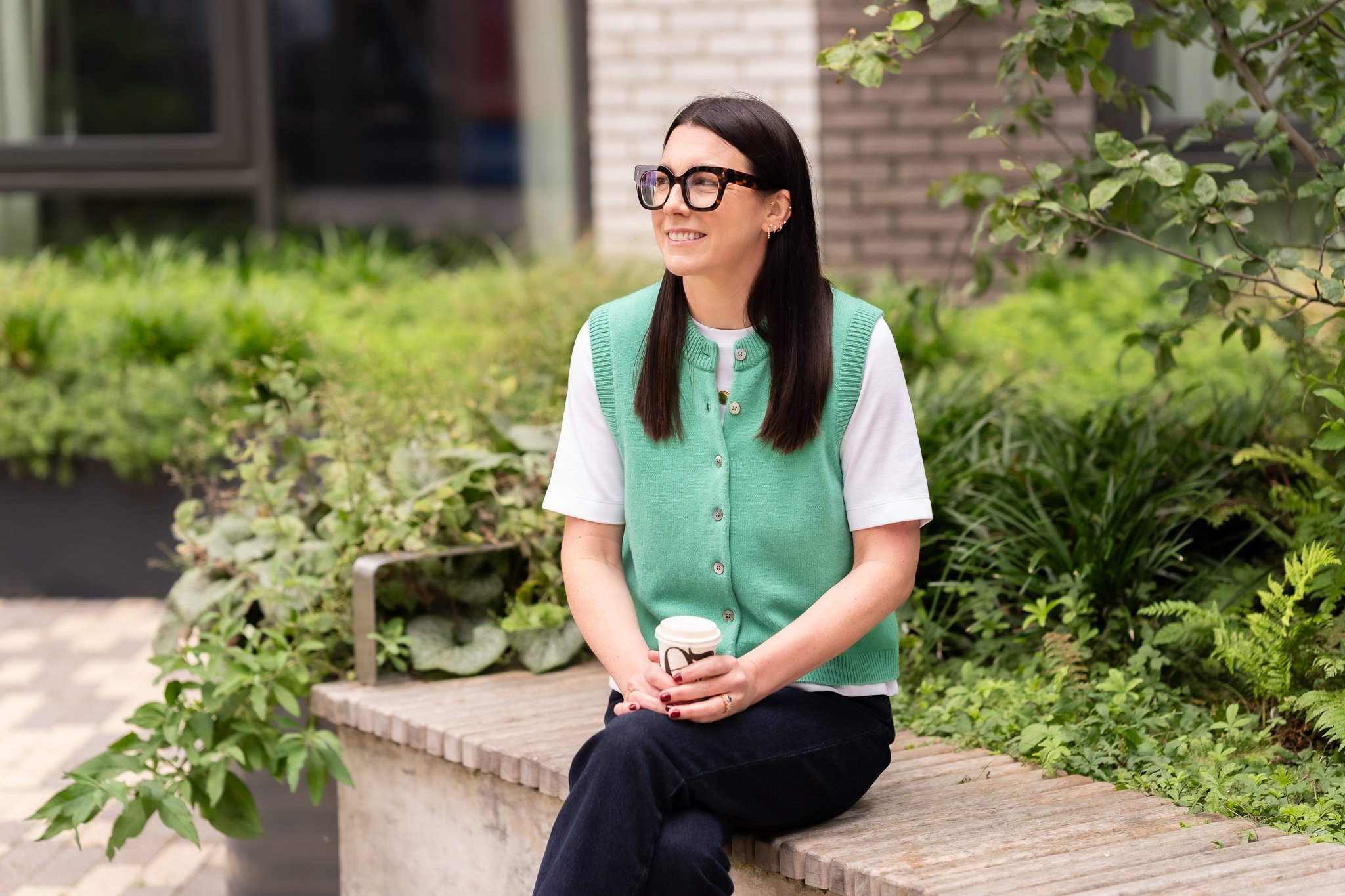 Harriet wearing a green top on a bench holding a coffee