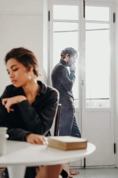 Man standing by window smoking and woman sitting at table looking the other way