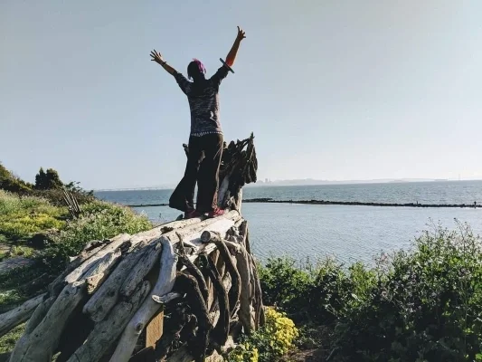 Person standing on hand built wooden structure with arms up overlooking body of water