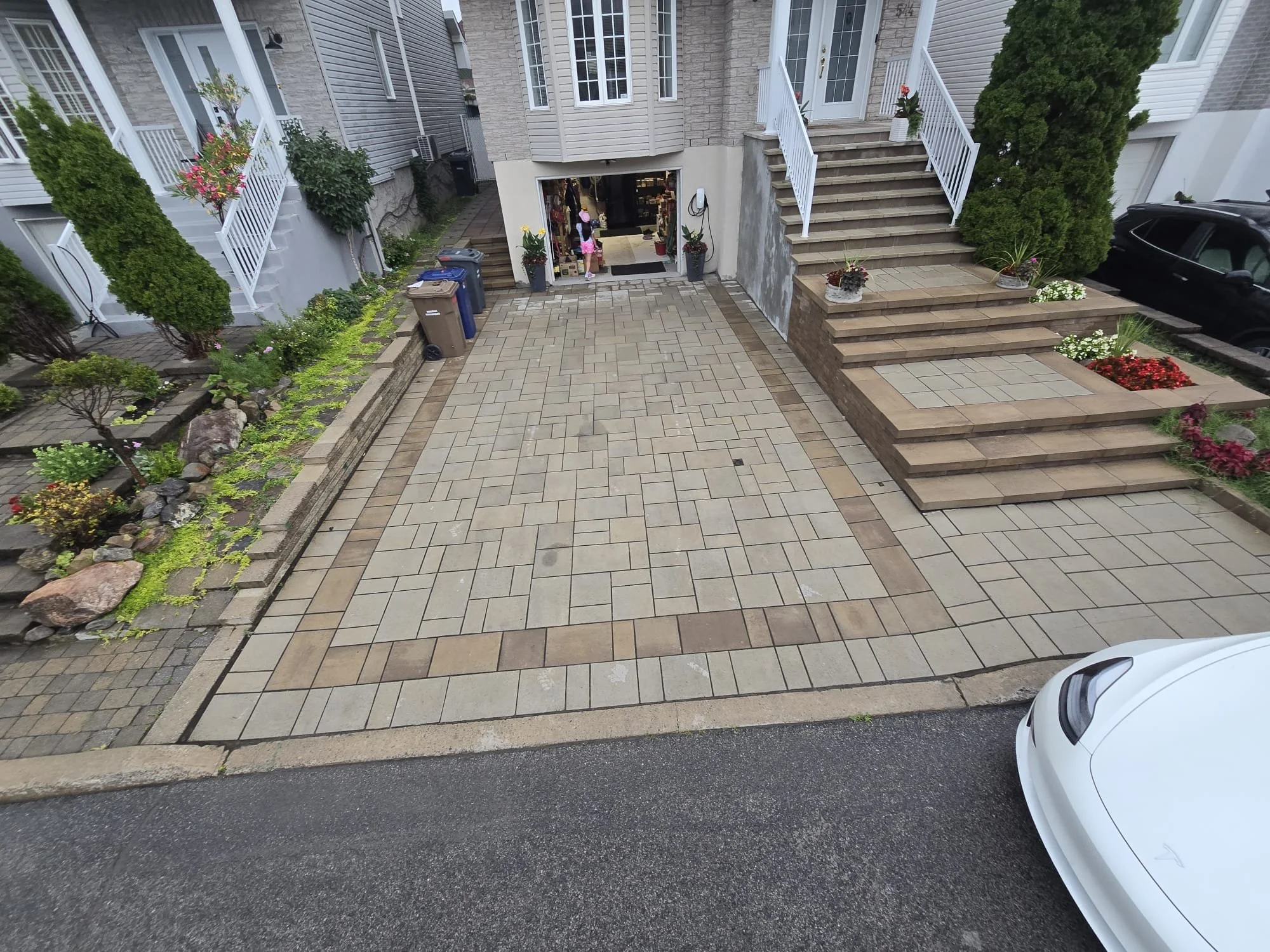 View of a residential driveway with decorative pavers, a staircase leading to the house entrance, and landscaped plants and flowers on both sides.
