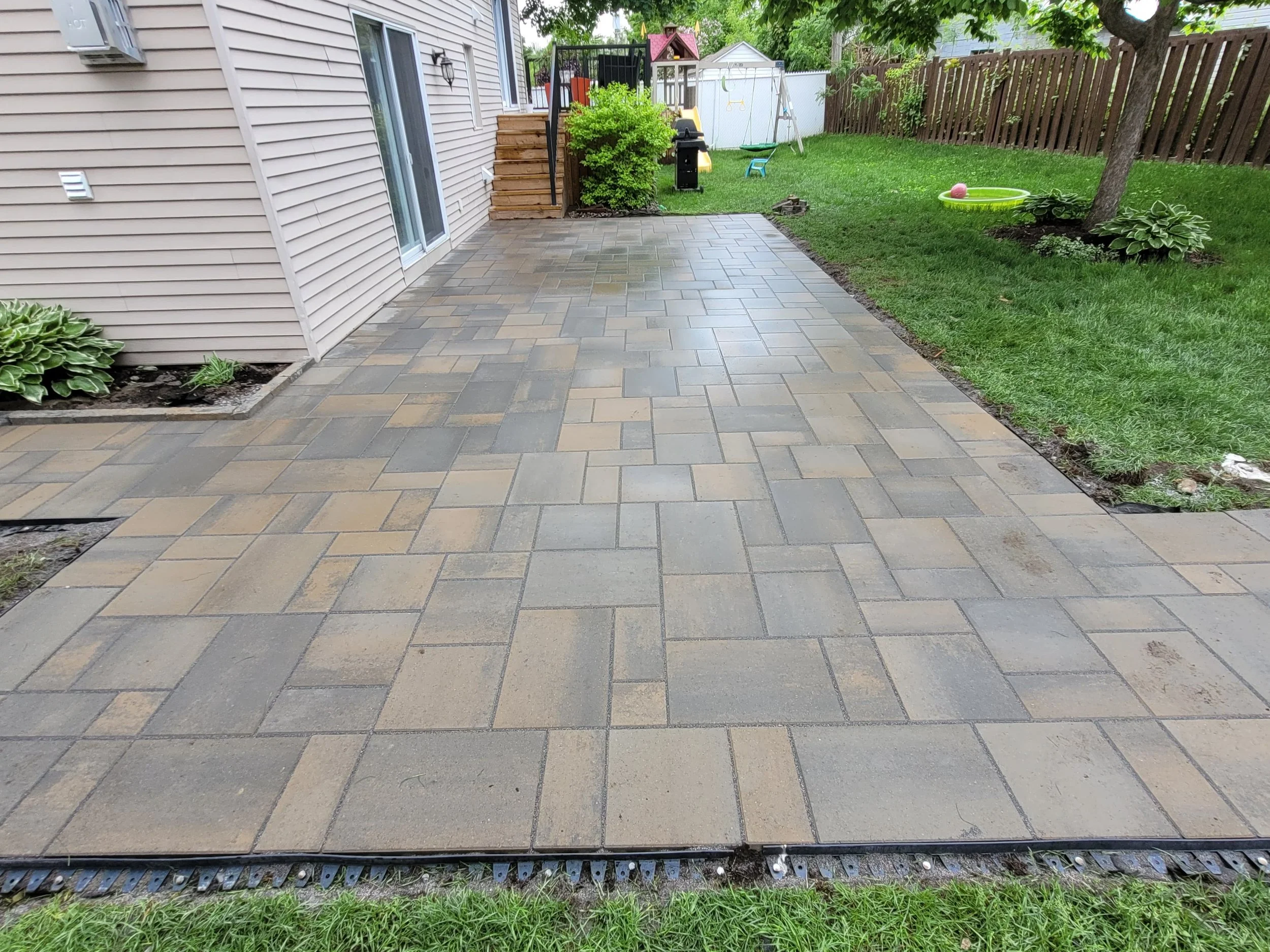 Backyard patio with newly installed paver stones, adjacent to a house with beige siding, with green grass and children’s play equipment in the yard.