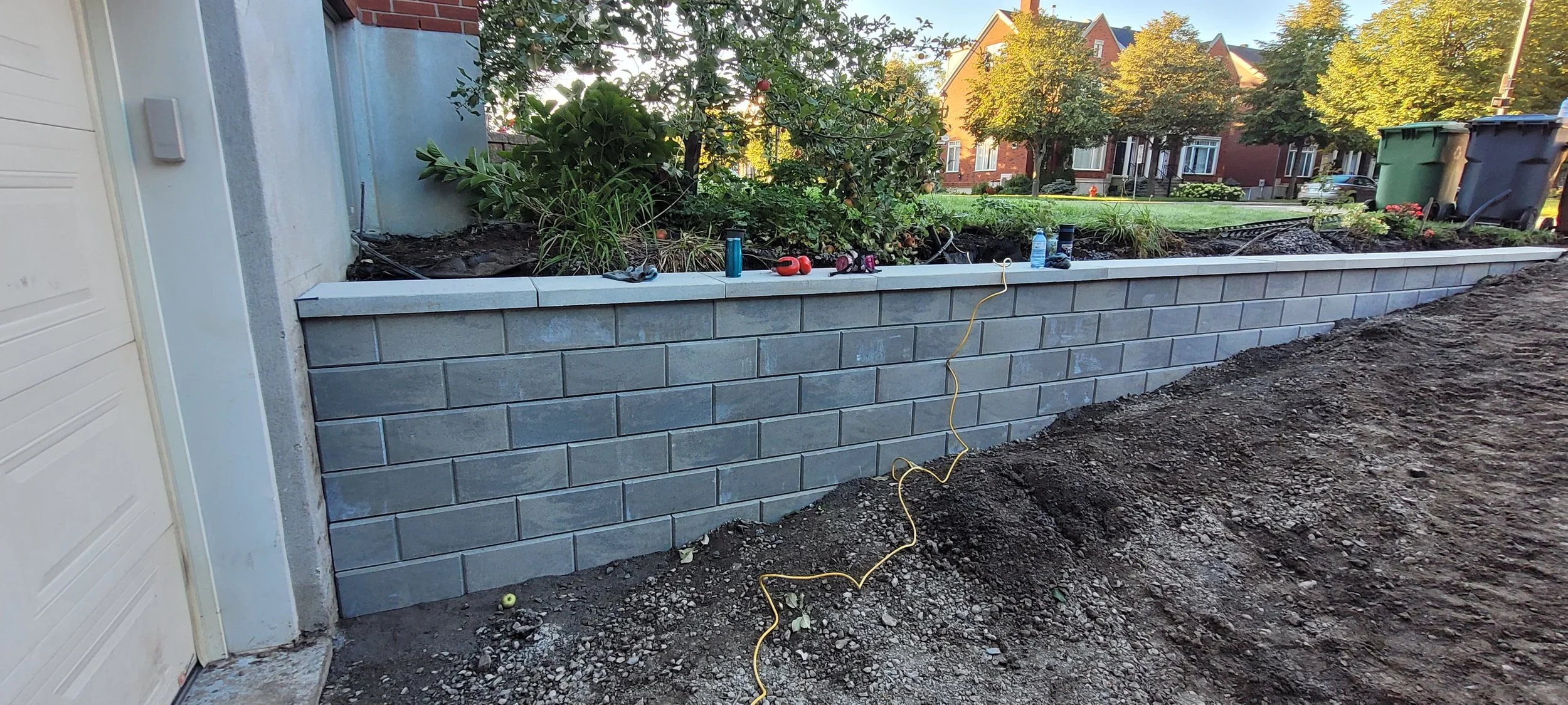 Construction site with a new gray brick retaining wall and landscaping work in progress, tools, a yellow extension cord, and neighboring houses in the background.