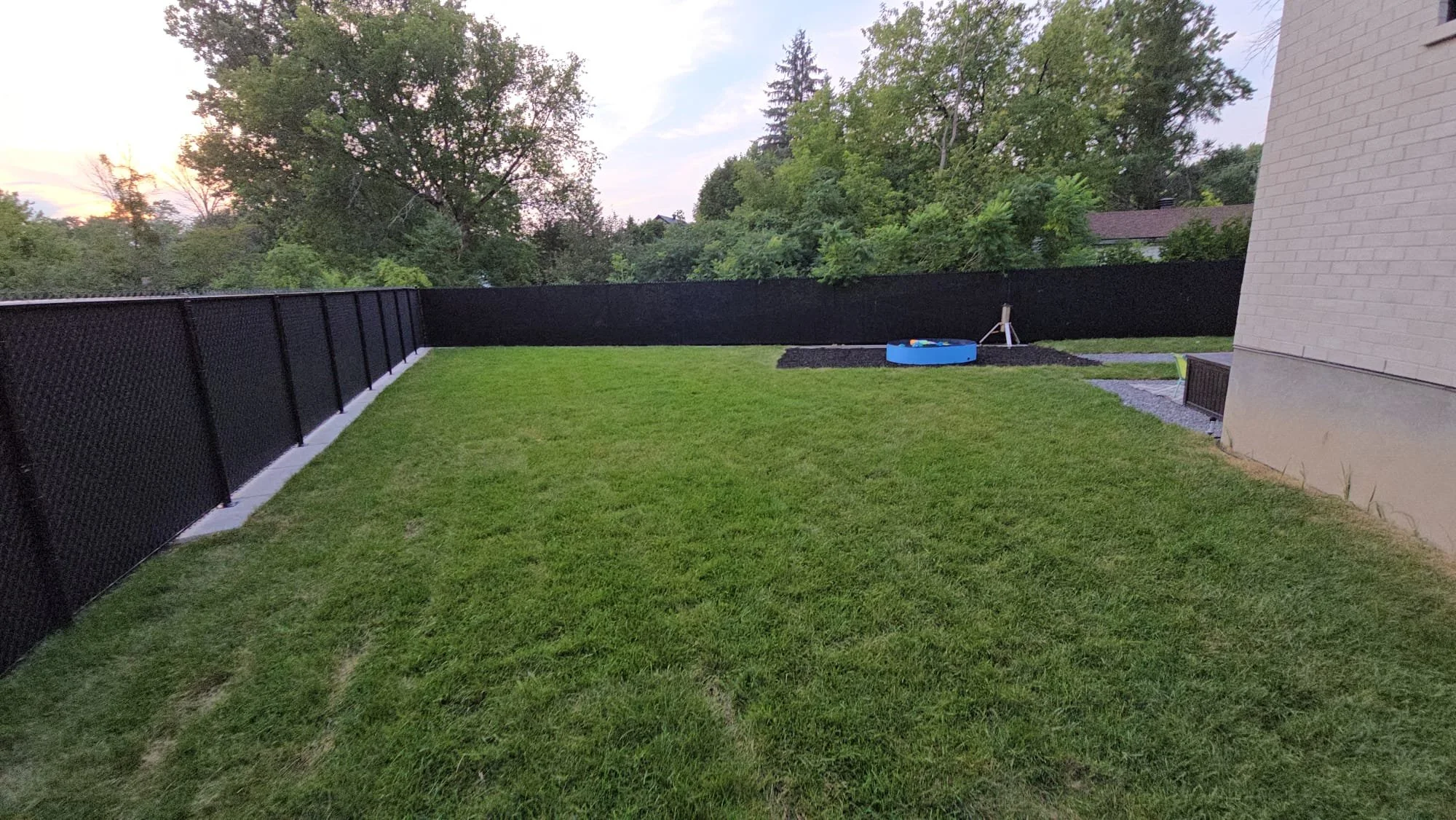 A backyard with a green lawn, black fence, some trees in the background, and a small blue plastic sandbox on a gravel area near the house.