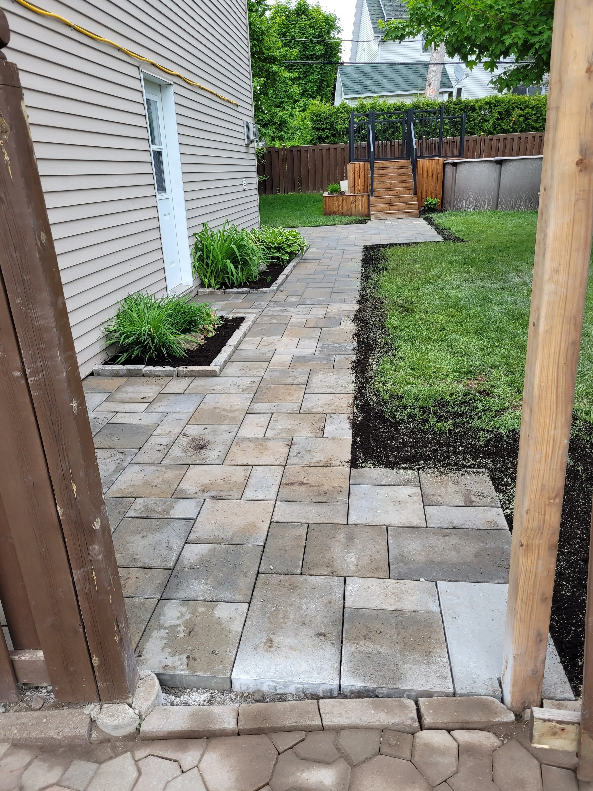 A backyard with a partially completed stone walkway, green grass, planted flowers, and a wooden deck with stairs in the background.