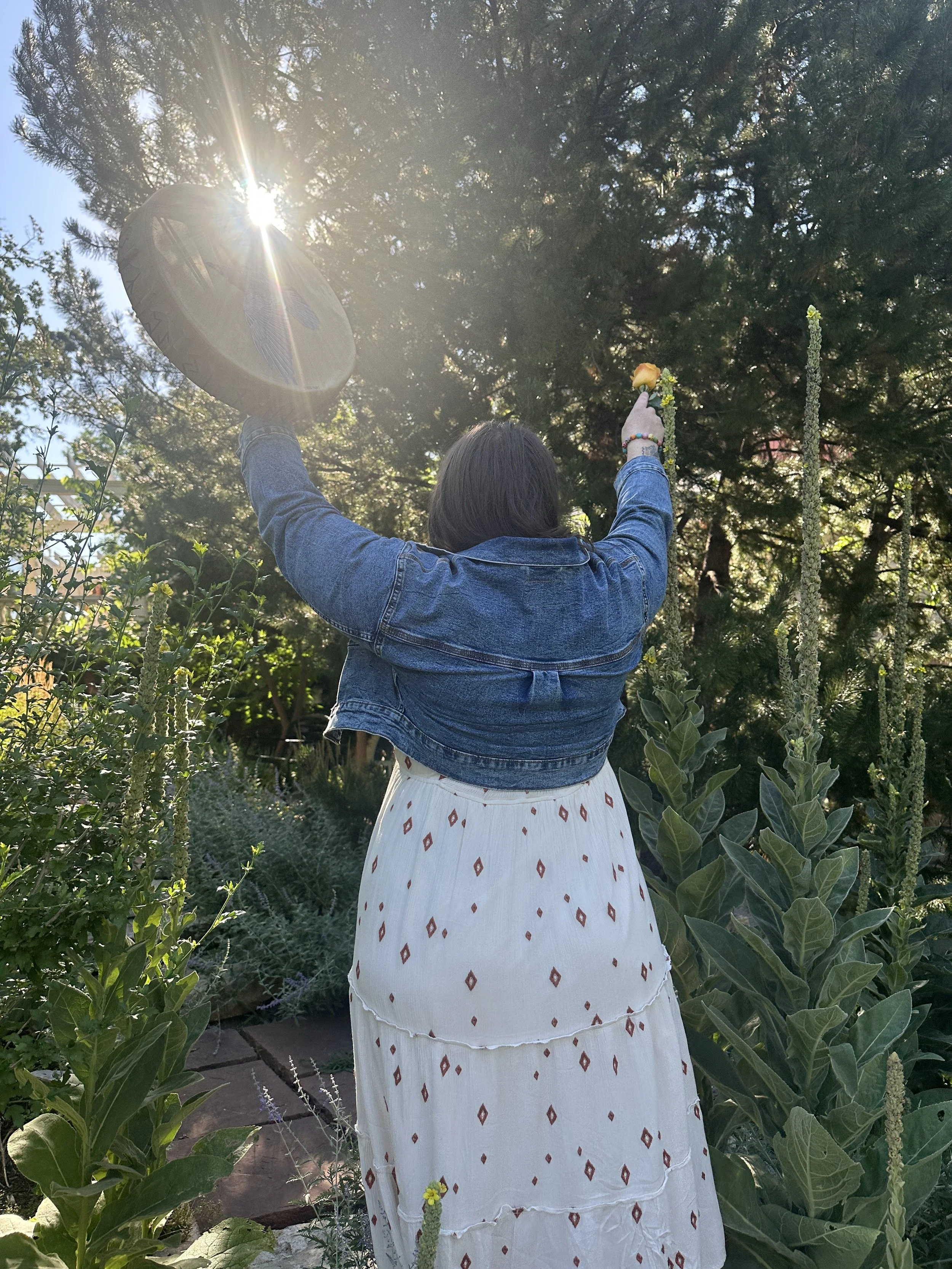 Isabel standing outdoors among green plants, facing away from the camera, holding a drum in one hand and a small flower in the other, with sunlight shining through the trees.