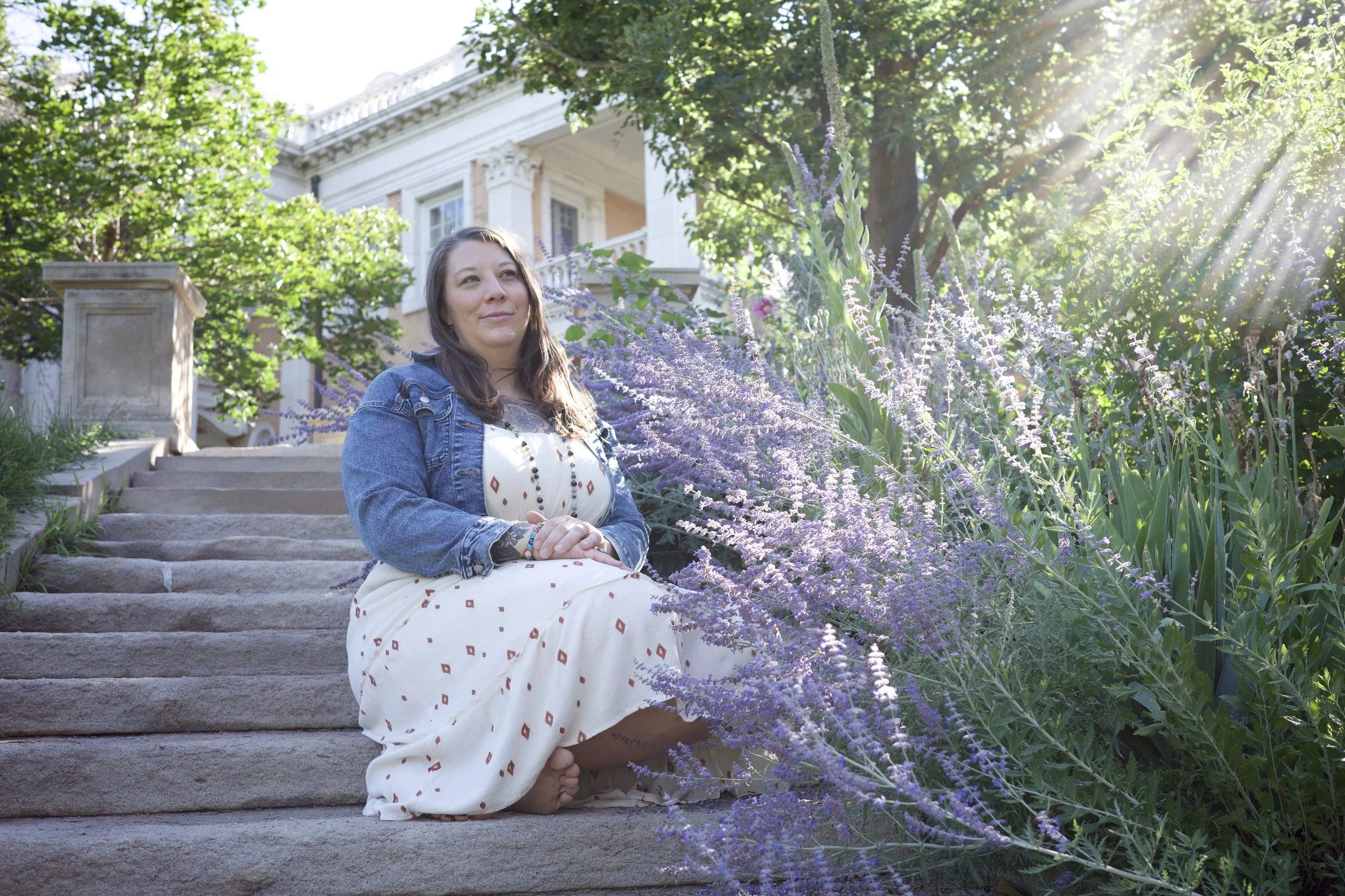Isabel sitting on stone steps outdoors, surrounded by russian sage, with sunlight filtering through trees and a house in the background.