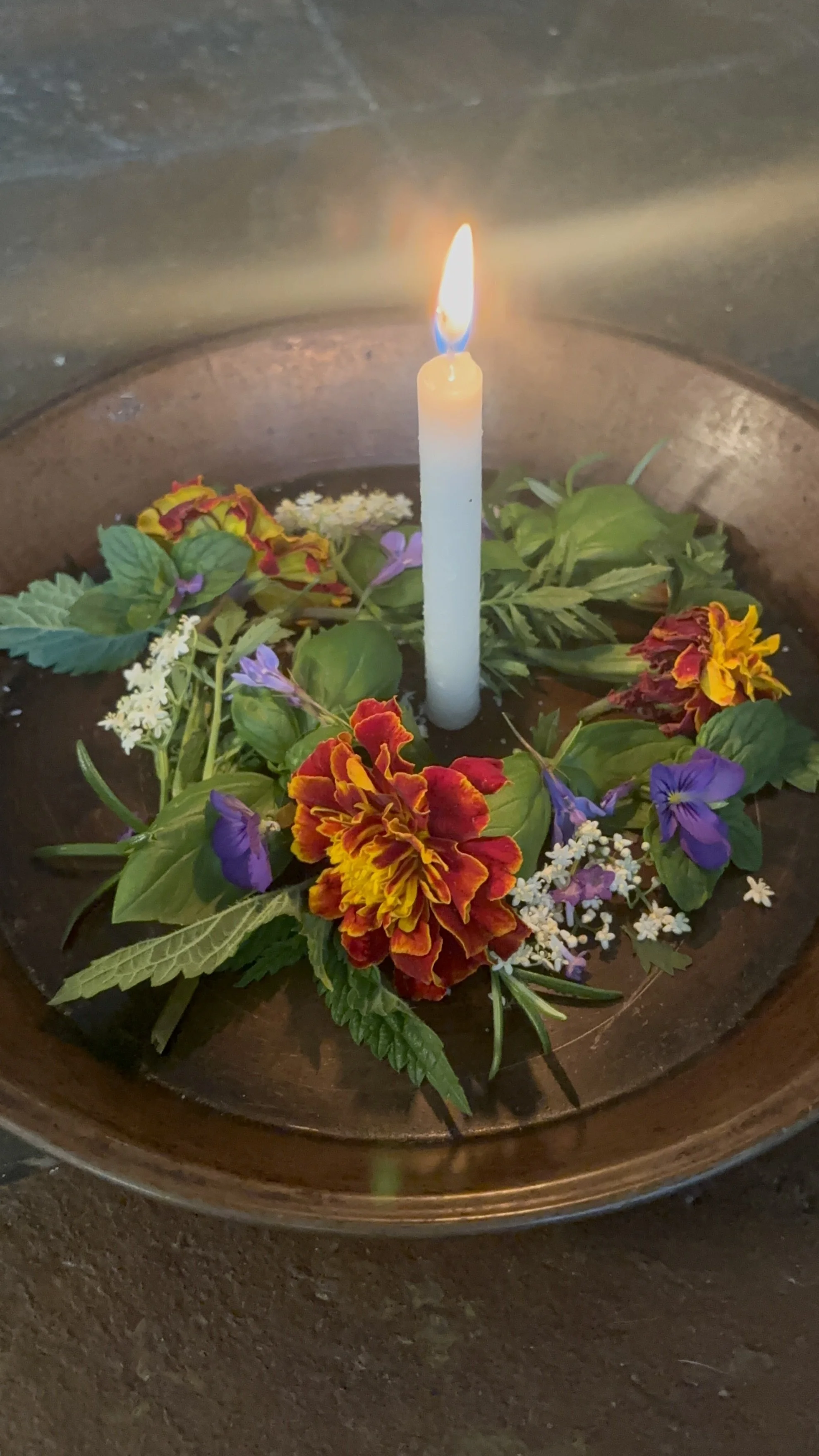 A white candle burning in the center of a floral arrangement with colorful flowers and green leaves in a copper basin.