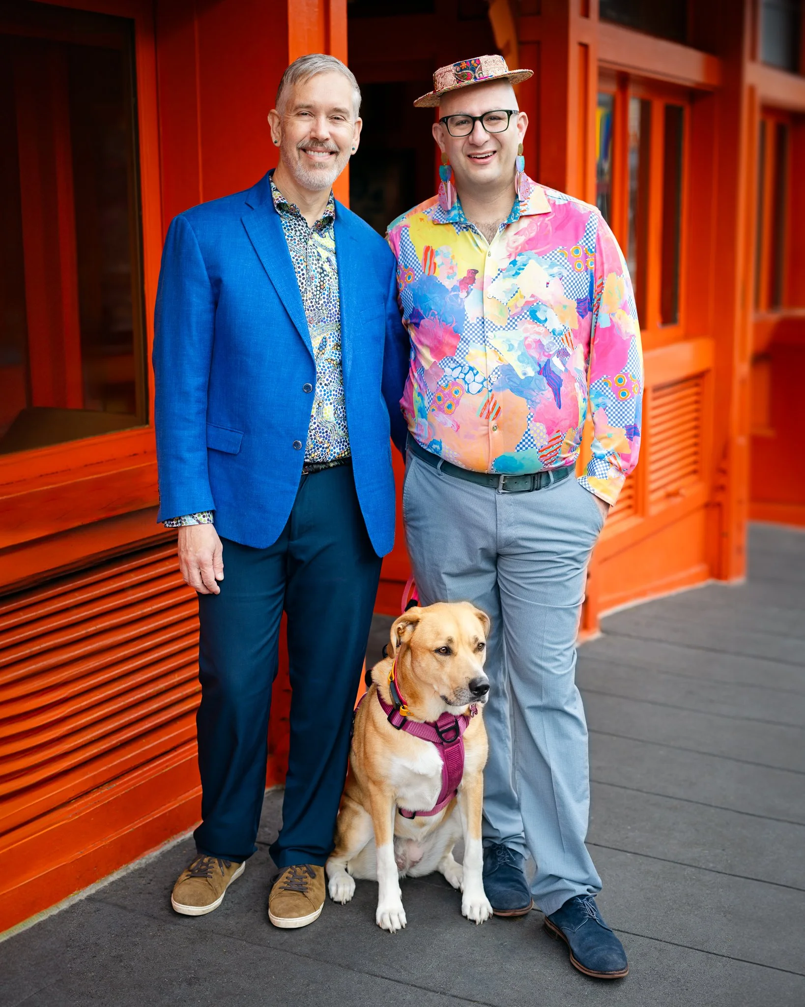 Kevin, Pumpkin, and Alexis standing in front of a local taqueria. Alexis is wearing a pasted-colored, patterned shirt and a hat. Kevin is wearing a blue coat and dark blue pants. Pumpkin is sitting between them.