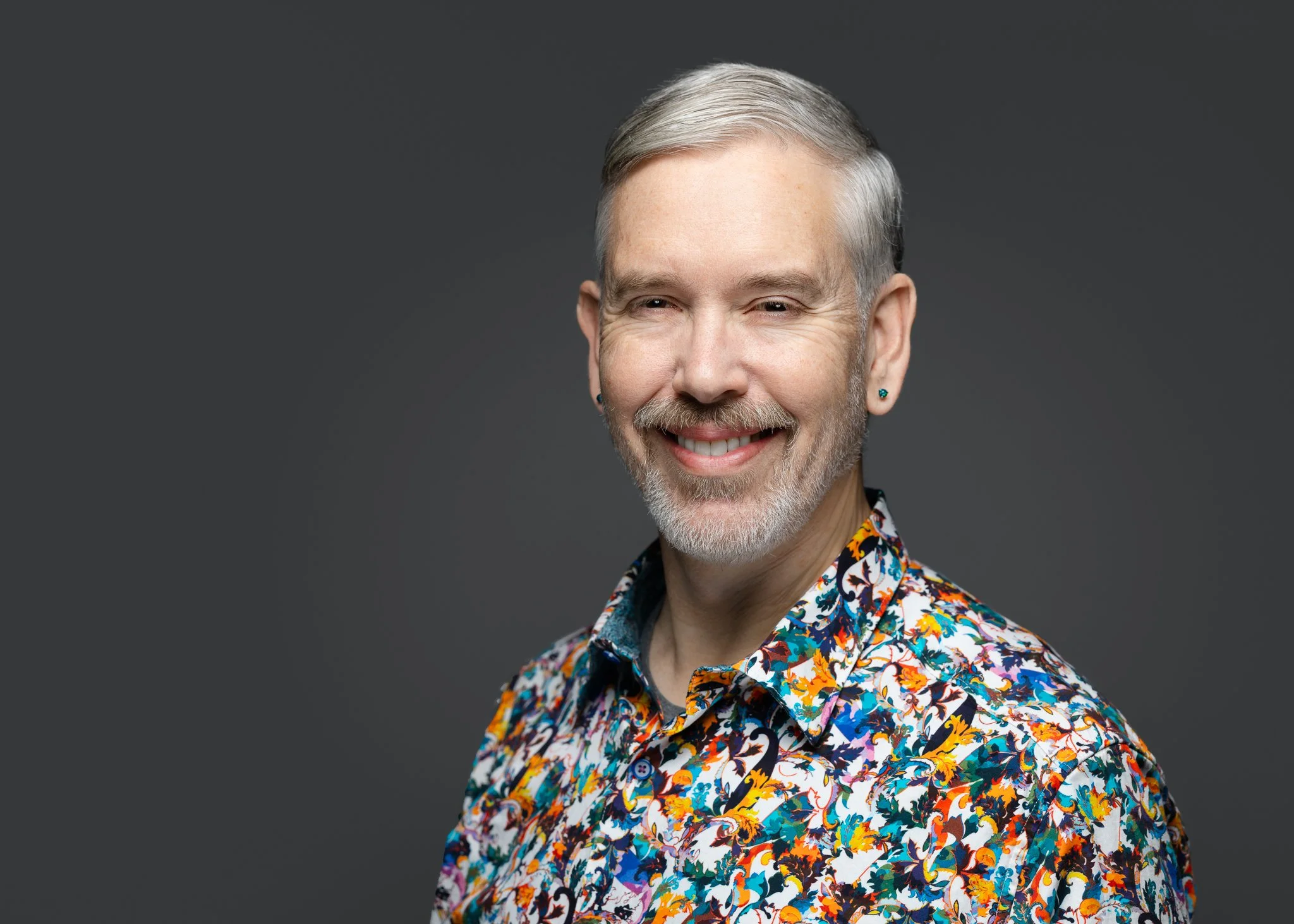 Portrait of Kevin, who has gray hair and beard. He is wearing a multi-colored, button-down shirt and giving some smile!