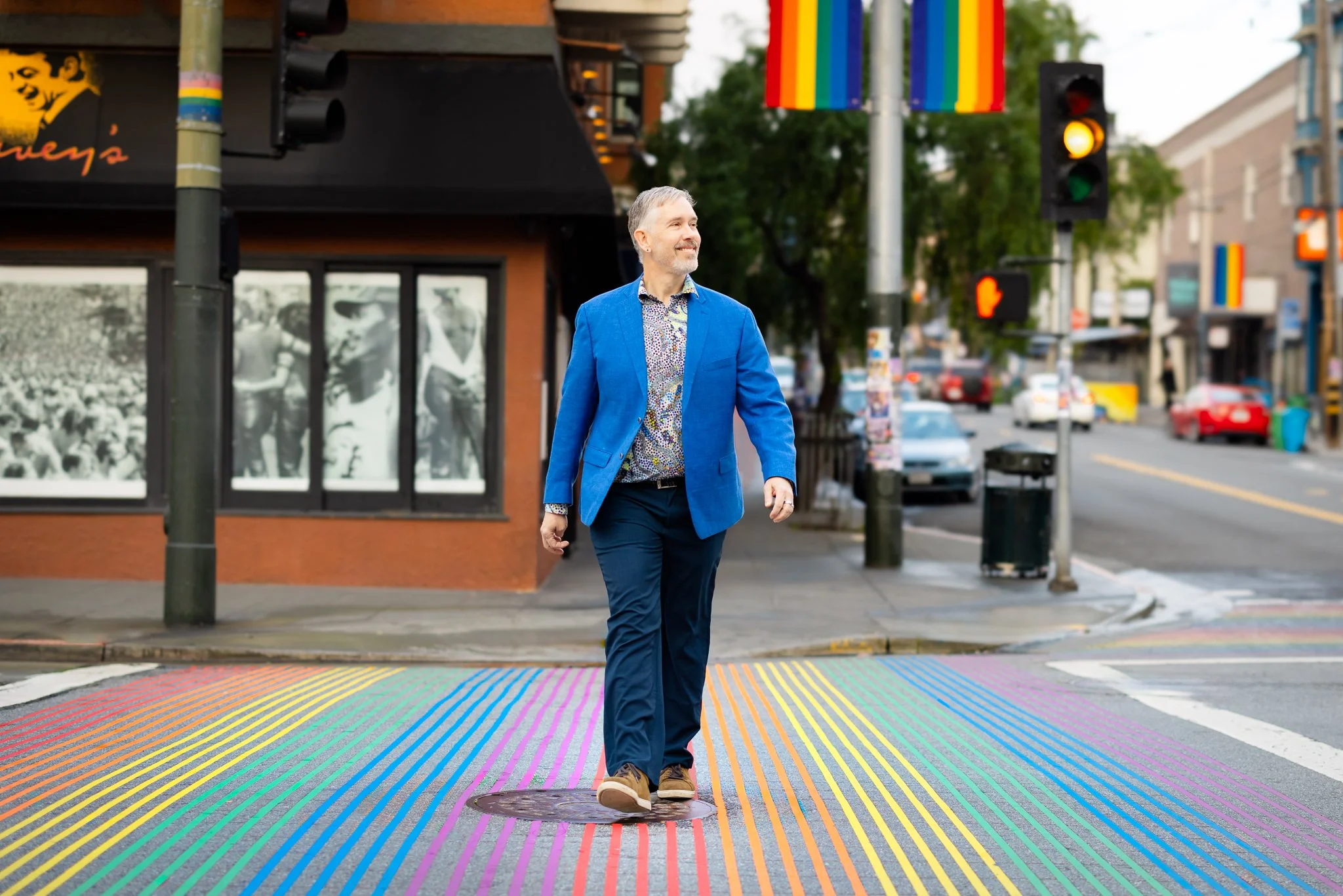 Kevin, wearing a blue coat over a variegated shirt, dark blue pants, and goldenrod shoes crossing Castro Street in the rainbow crosswalk. He is looking off to the left and smiling.