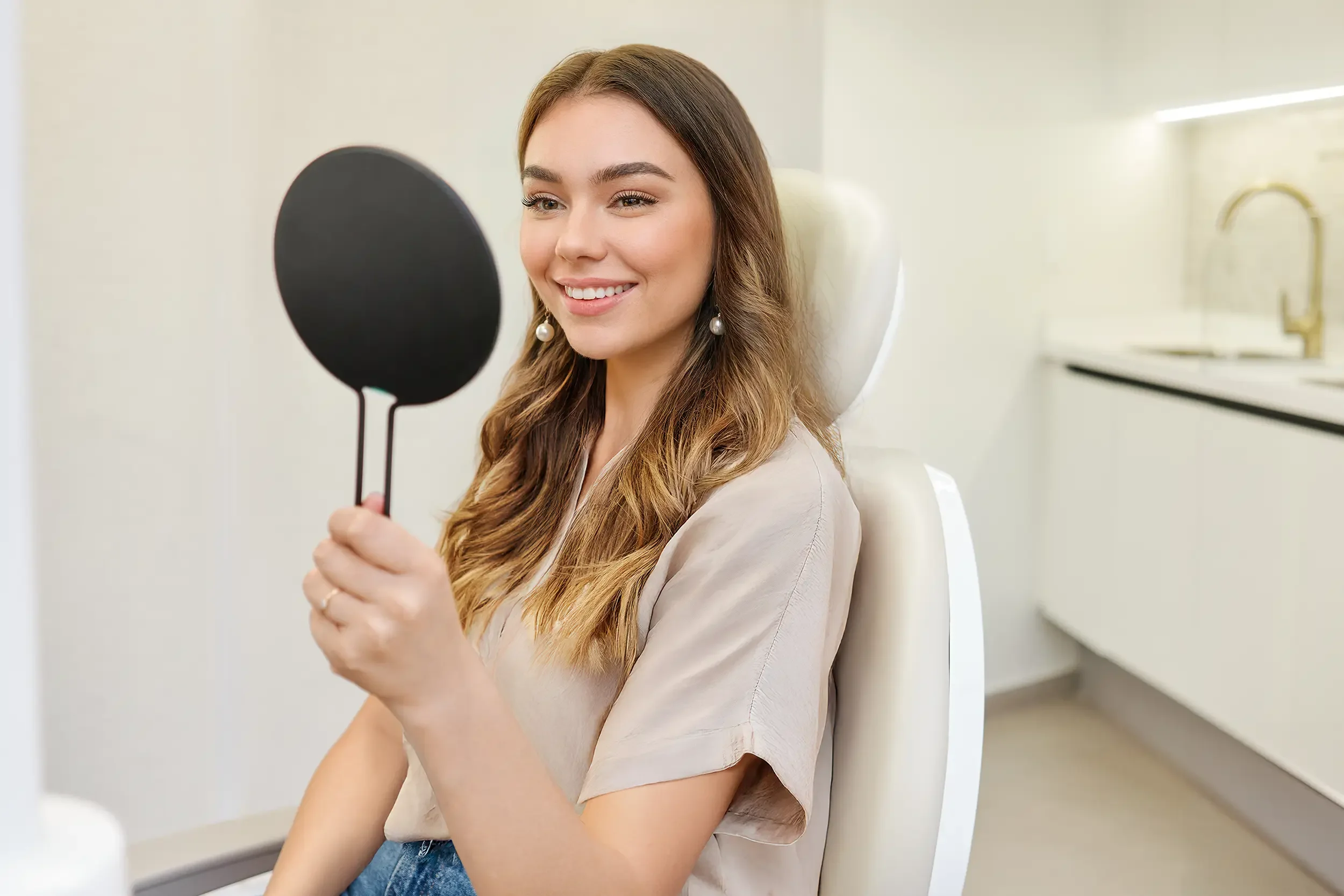 Joven mujer sonríe al espejo después de blanqueamiento dental en Clínica Aires.