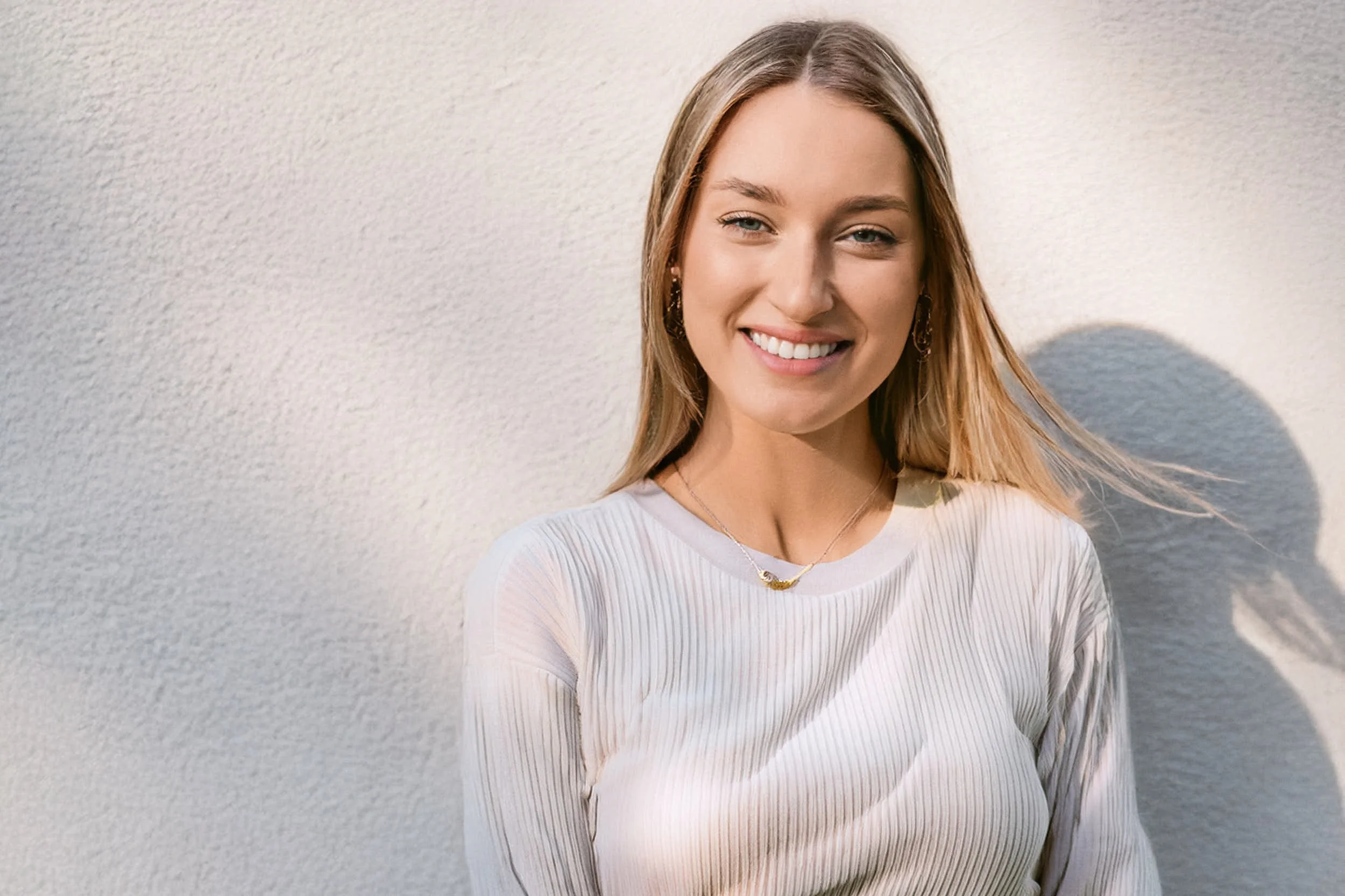 Mujer joven con blusa blanca y pendientes, sonriendo, con fondo de pared blanca.