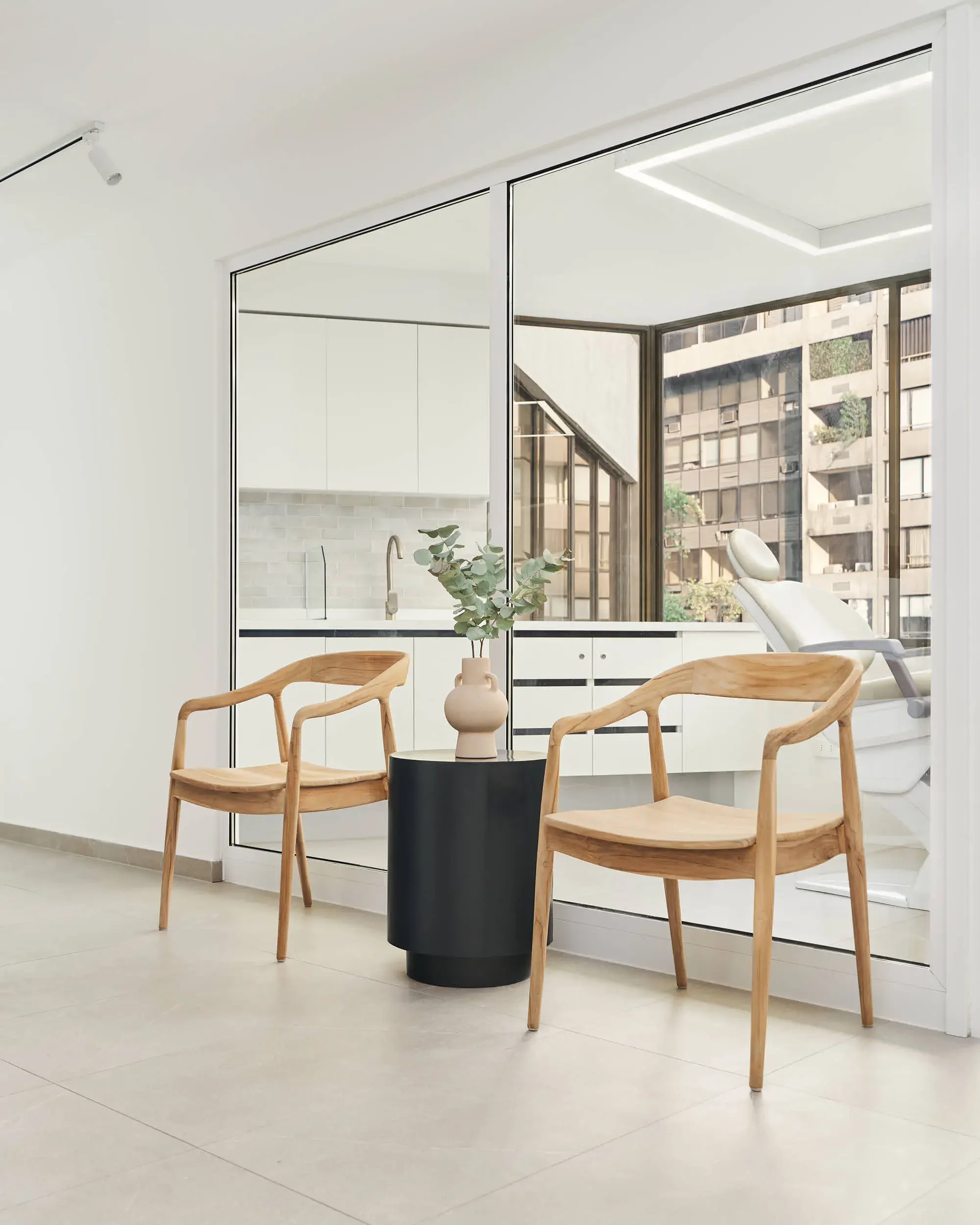 Sala de espera moderna de Clínica Aires. con dos sillas de madera, una mesa negra con un florero, suelo de azulejos y grandes ventanales que muestran un edificio. Al fondo, una sala con equipo médico.