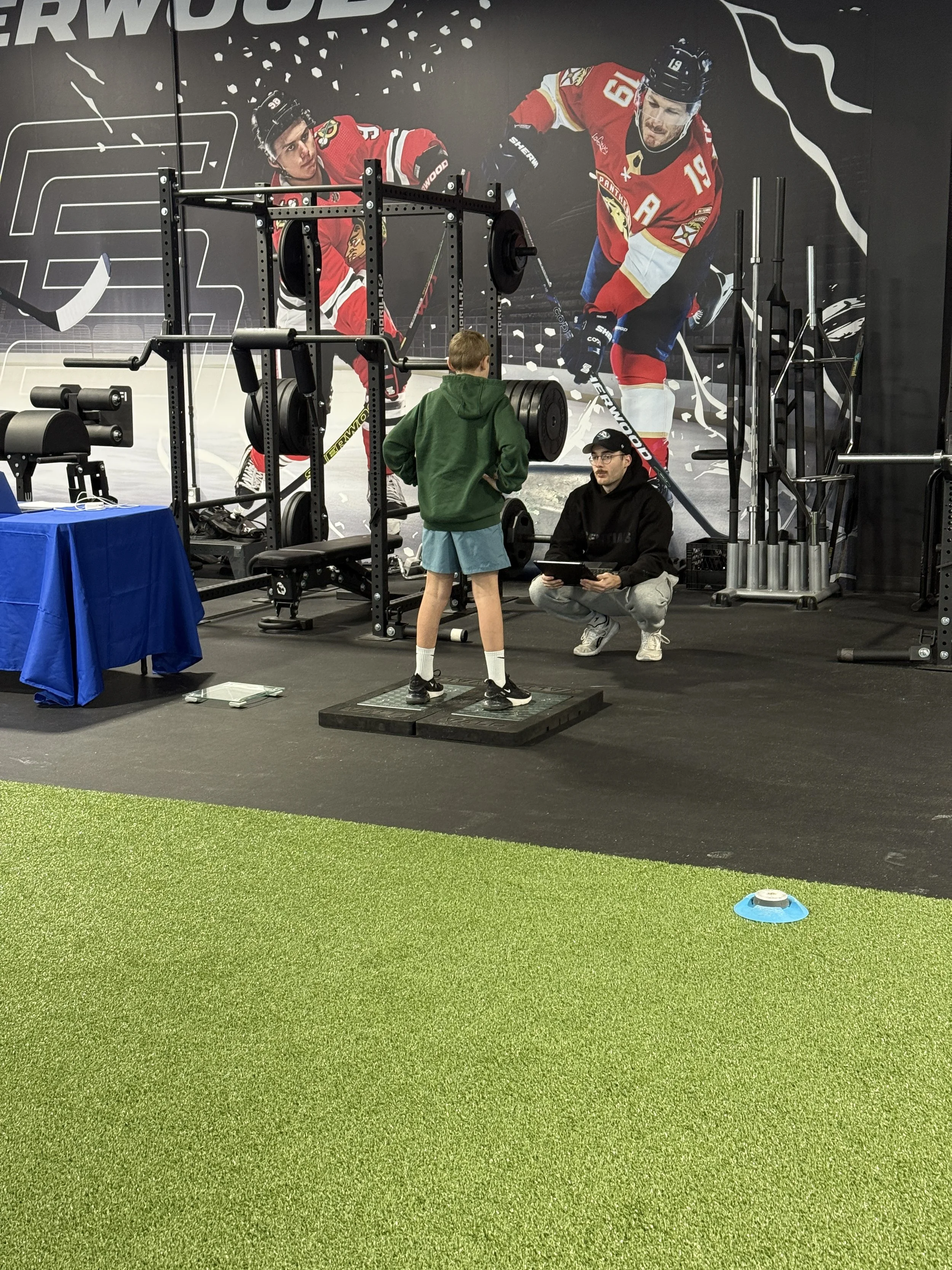 Children and adults engaging in a training session at a gym. Kids are practicing with medicine balls and cones on an artificial turf. Large exercise balls are visible on shelves at the back, along with various gym equipment and free weights.