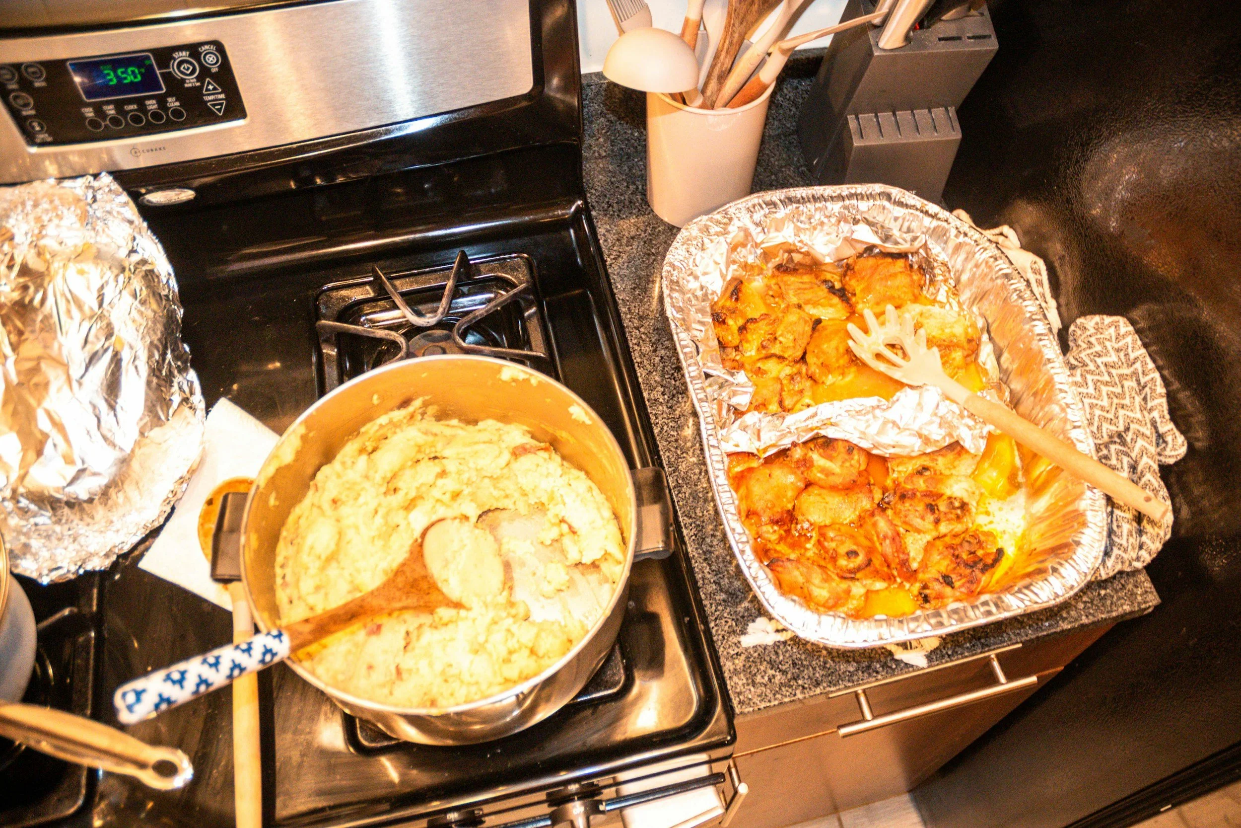 Pot of mashed potatoes on stove, aluminum tray with cooked chicken in sauce, and wrapped foil dish on kitchen counter.