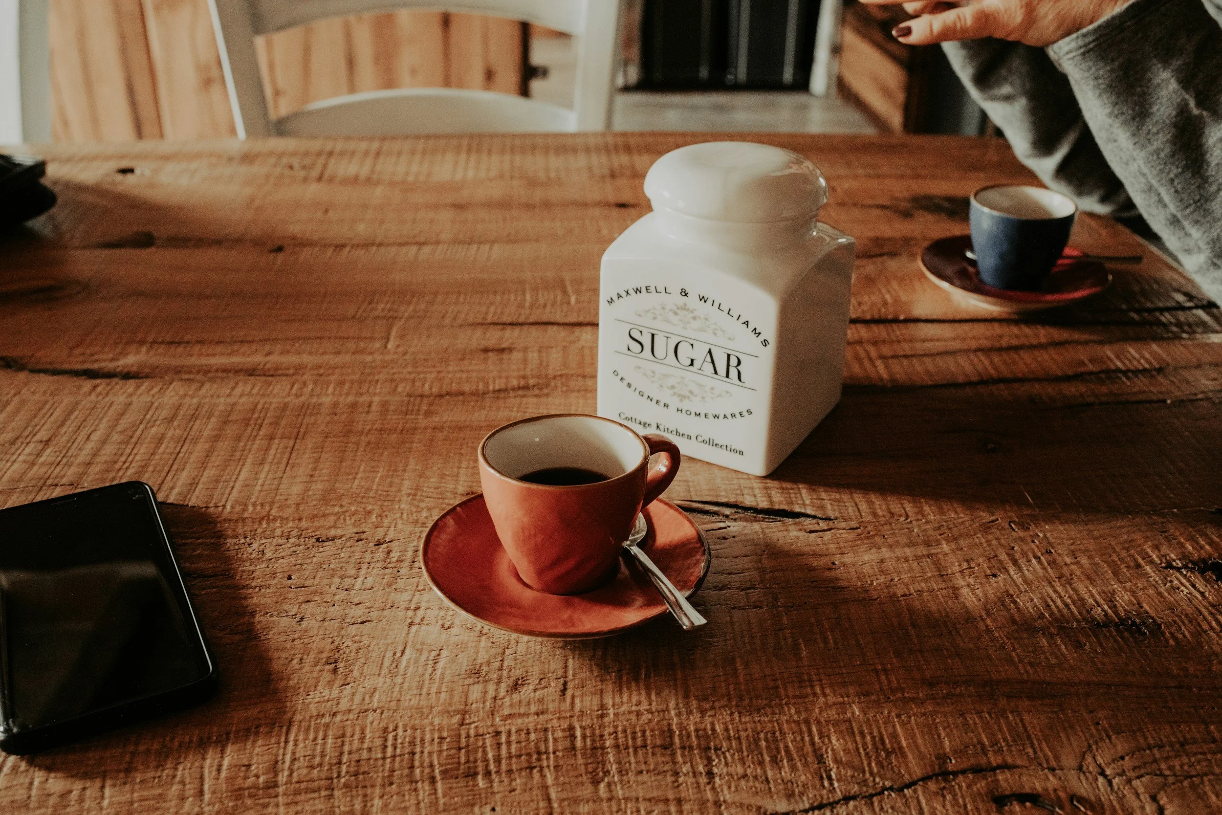 A rustic wooden table with a red cup of coffee on a matching saucer, a spoon, a white sugar container labeled 'Sugar,' and two additional cups in the background, one blue and one cut off.