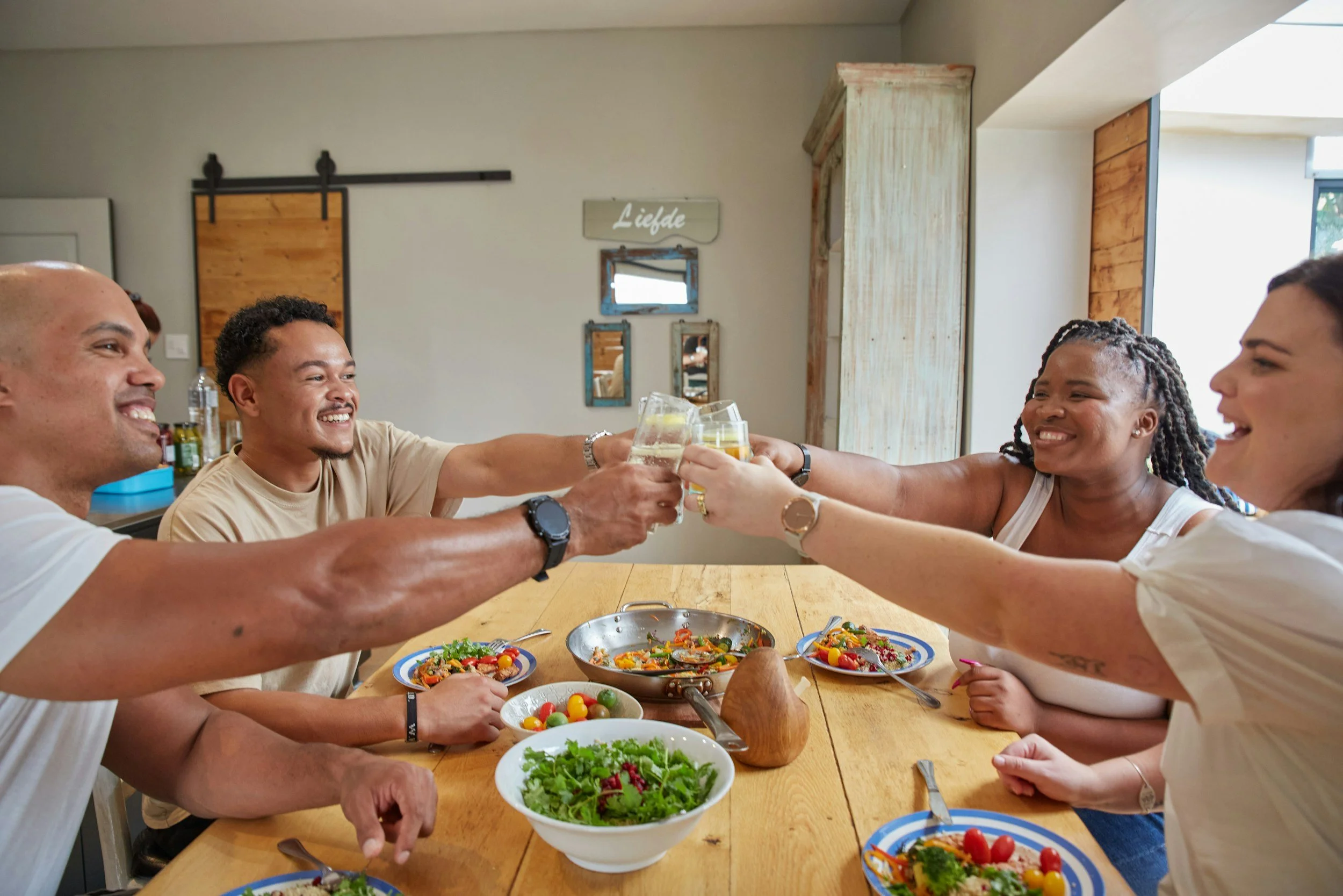 Five friends sitting around a wooden table, toasting with glasses of white wine, with plates of salad and food on the table in a bright, cozy dining room.
