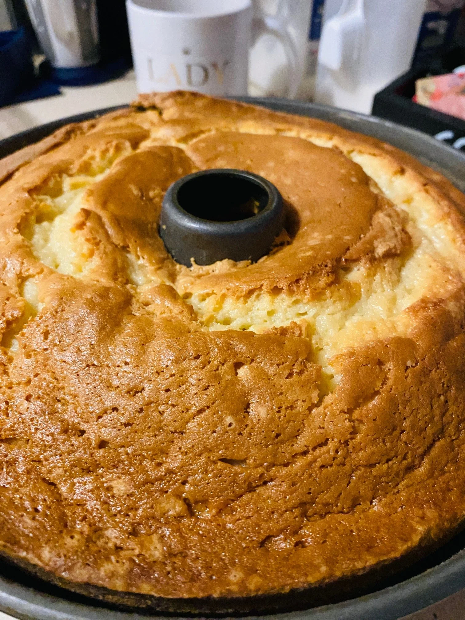 A close-up of a freshly baked, golden brown angel food cake with a black cake mold in the center, placed on a turntable in a kitchen.