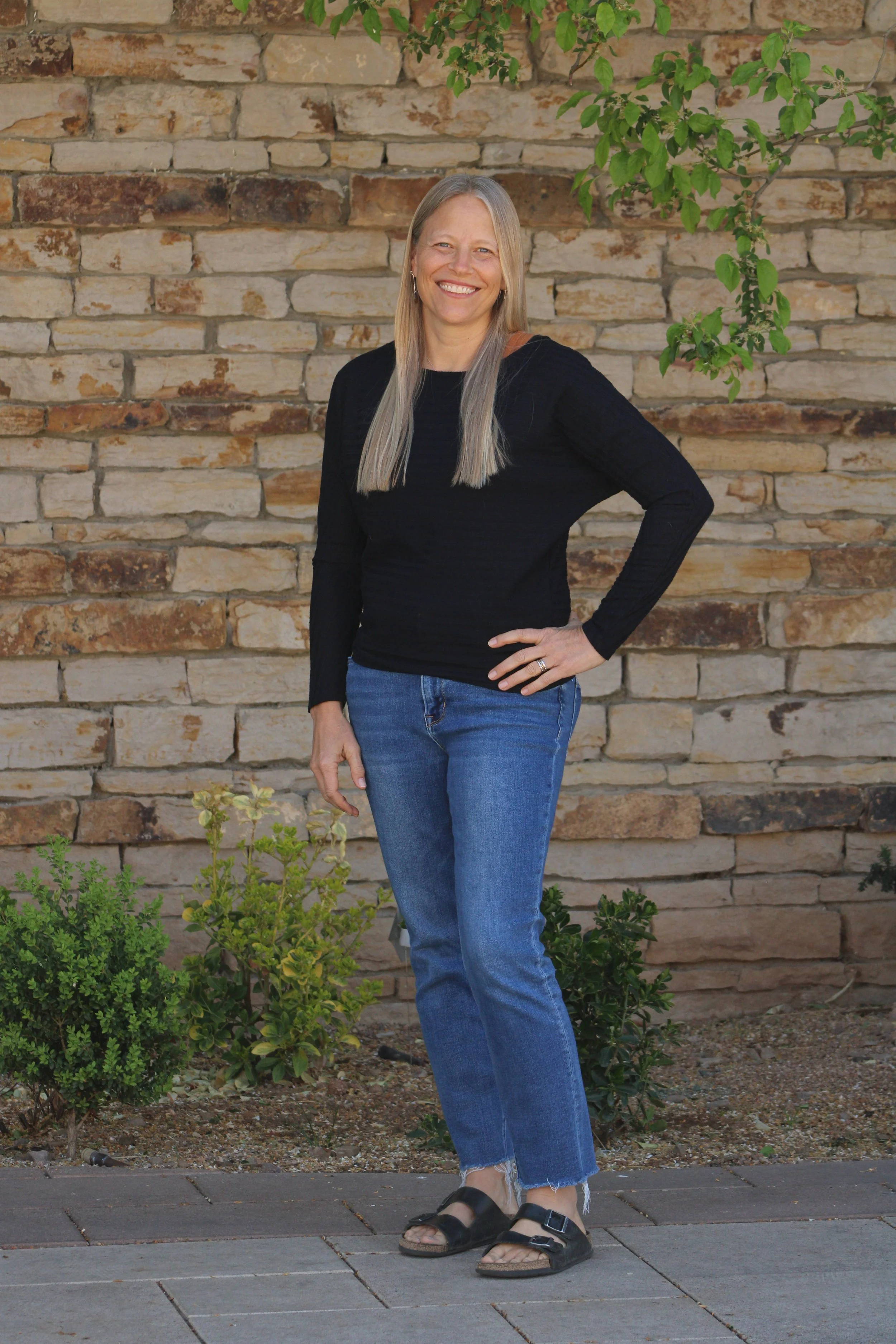 A woman stands outdoors in front of a stone wall with green plants, smiling at the camera. She has long, straight blonde hair and is wearing a black long sleeve shirt, blue jeans, and black sandals.