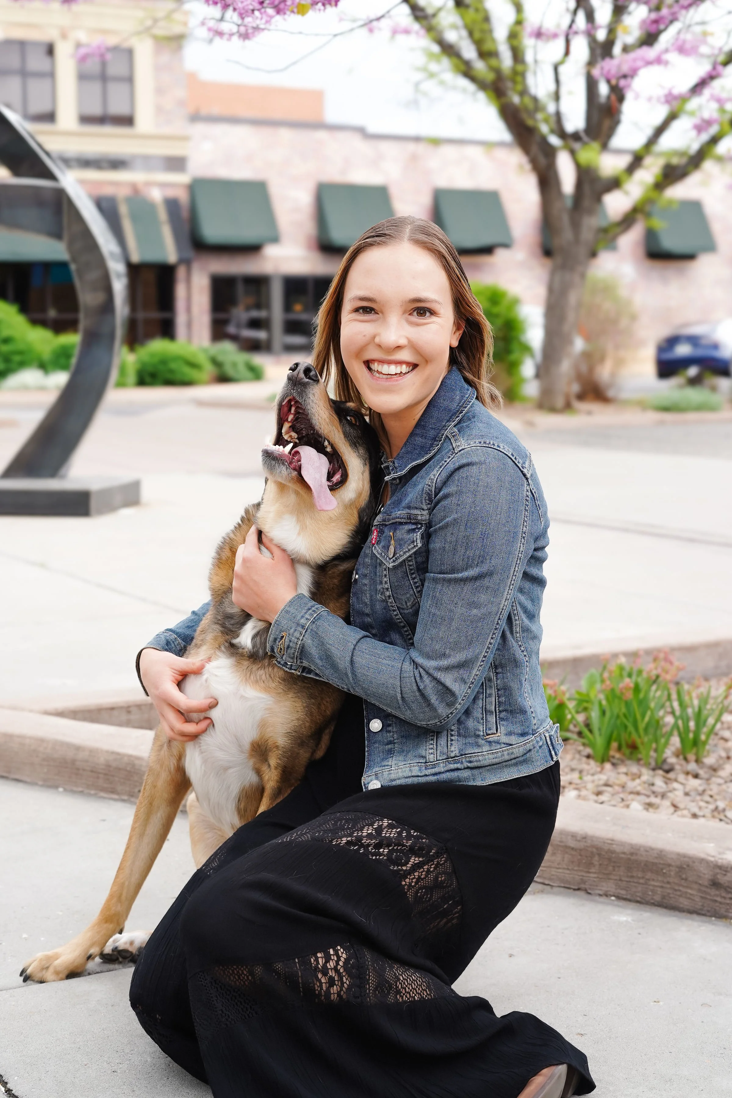 A woman smiling and holding a large dog, kneeling on a sidewalk in a city park, with a tree blooming in the background.