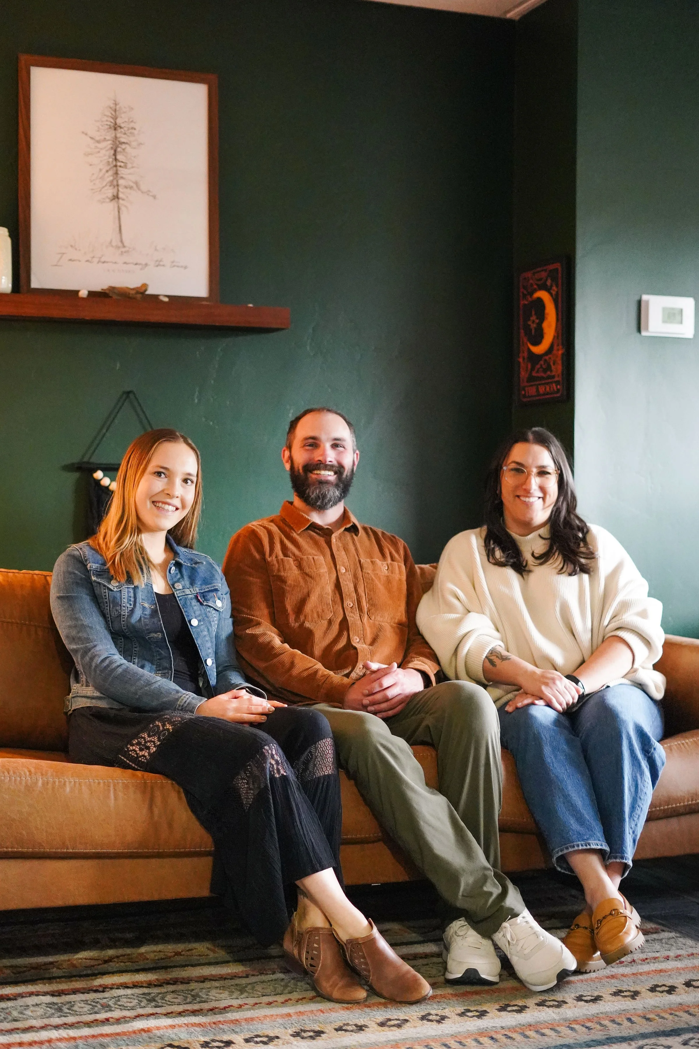 Three friends sitting on a brown sofa in a cozy room with dark green walls. The woman on the left has red hair, wearing a denim jacket and black lace pants. The man in the middle has a beard, wearing a brown shirt and khaki pants. The woman on the right has dark hair, glasses, a white sweater, and ripped jeans. They are smiling at the camera.