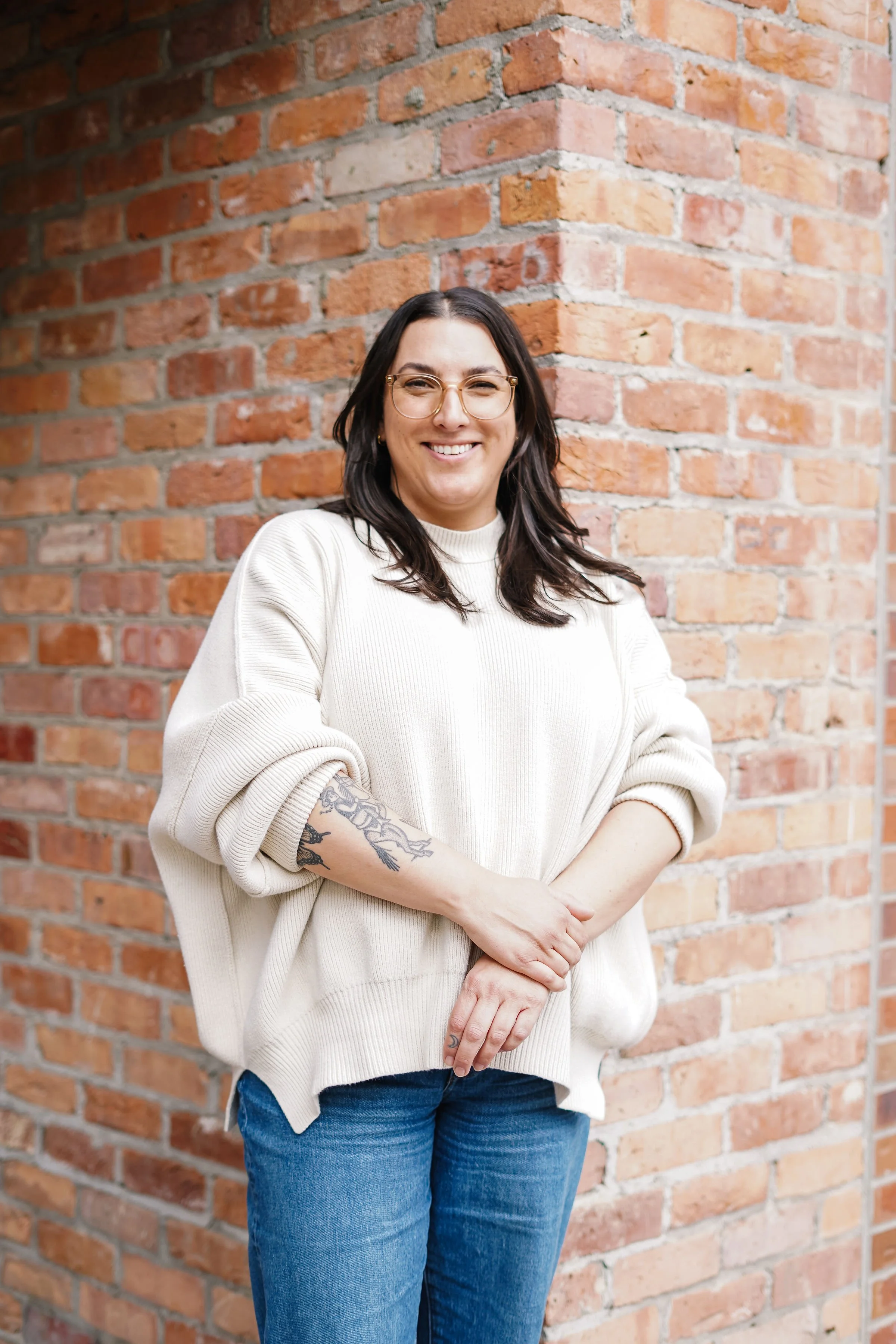 A woman with dark hair, glasses, and tattoos on her arm, smiling with arms crossed, standing in front of a brick wall.