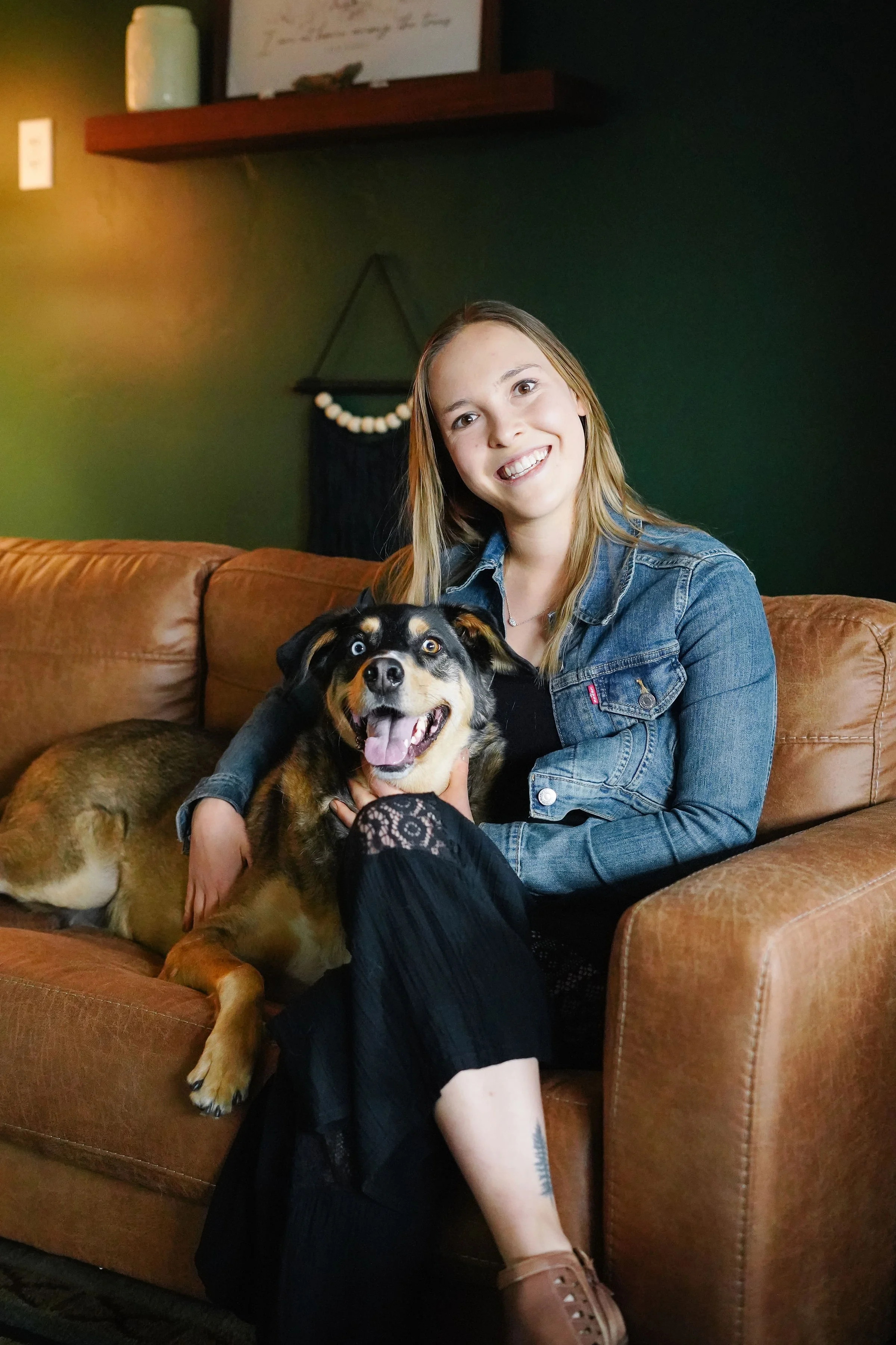 A young woman sitting on a brown leather couch with two dogs, one walking and another sitting on her lap. She is smiling and wearing a denim jacket and black lace dress.