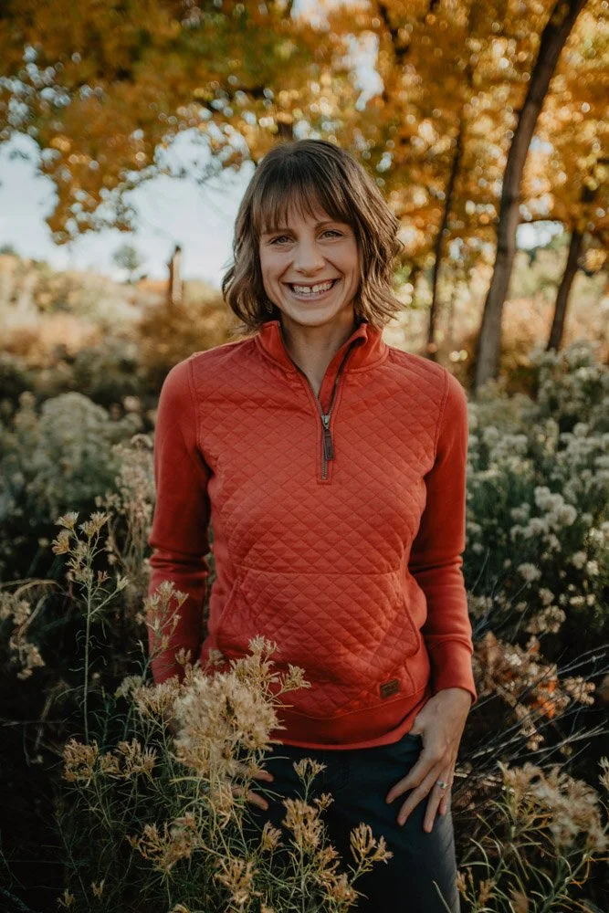 A smiling woman with short brown hair in a red quilted jacket standing outdoors among trees with autumn leaves and flowers.