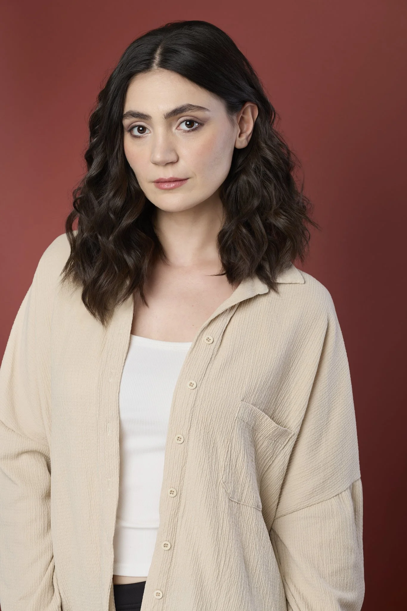 A young woman with shoulder-length wavy dark brown hair, wearing a beige oversized shirt over a white top, standing against a reddish-brown background.