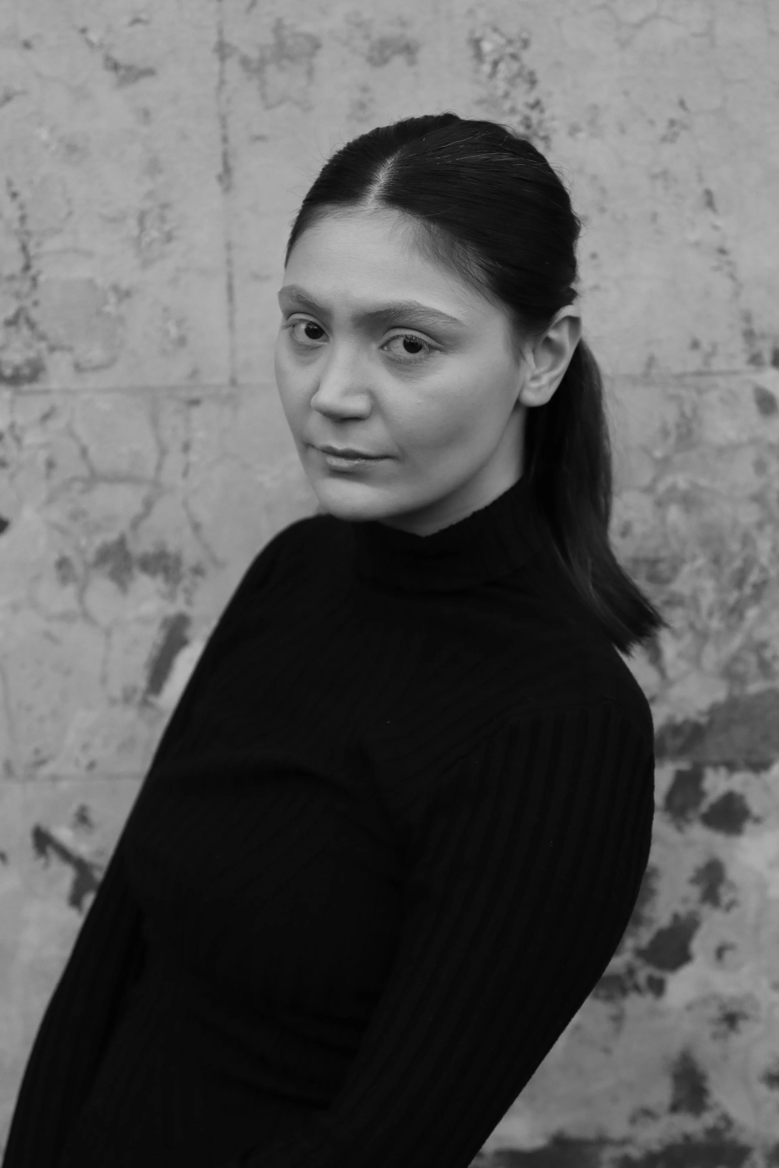 Black and white portrait of a woman with dark hair in a ponytail, wearing a black turtleneck sweater, standing against a textured wall.