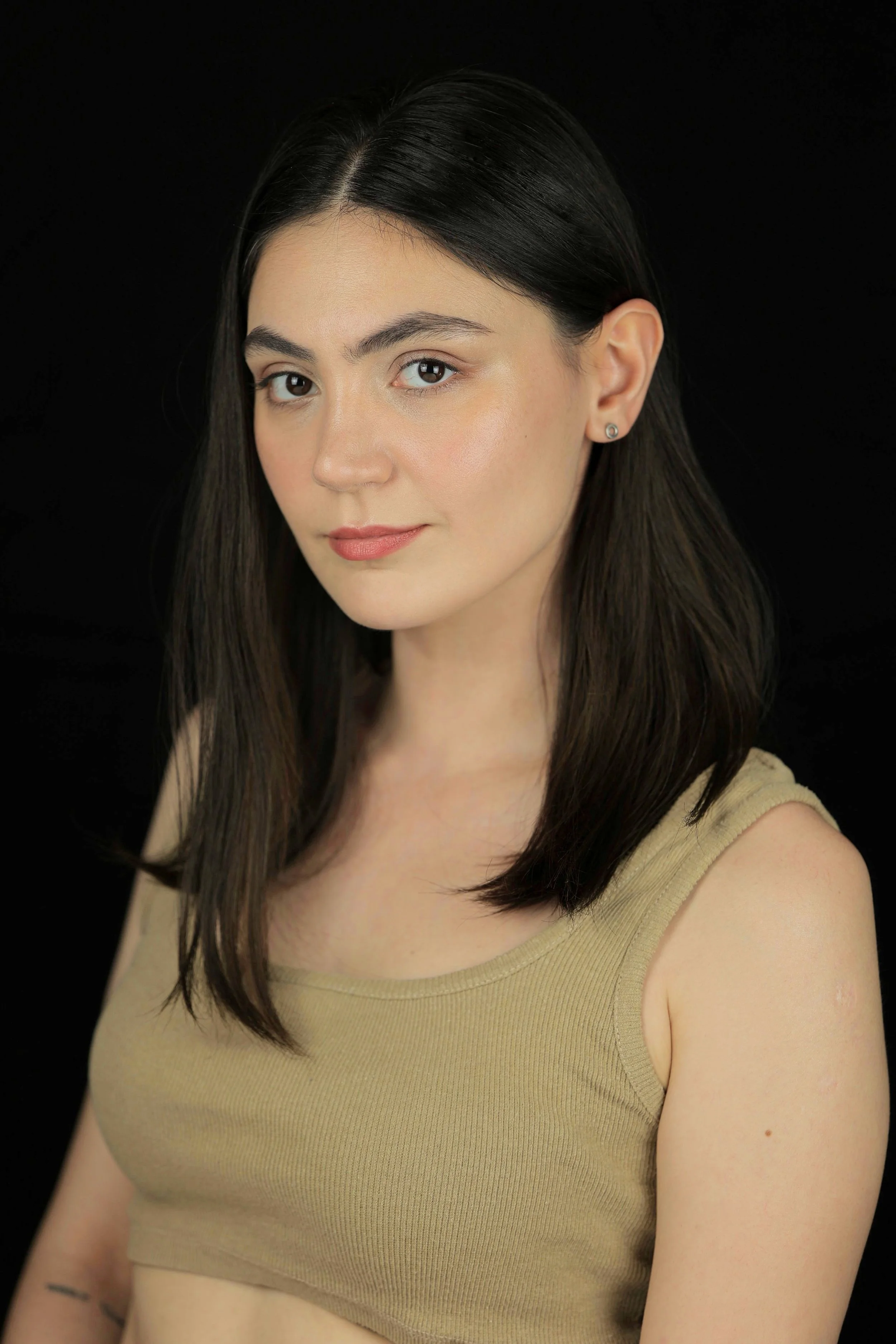Portrait of a young woman with straight dark hair, wearing a beige sleeveless top, against a black background.