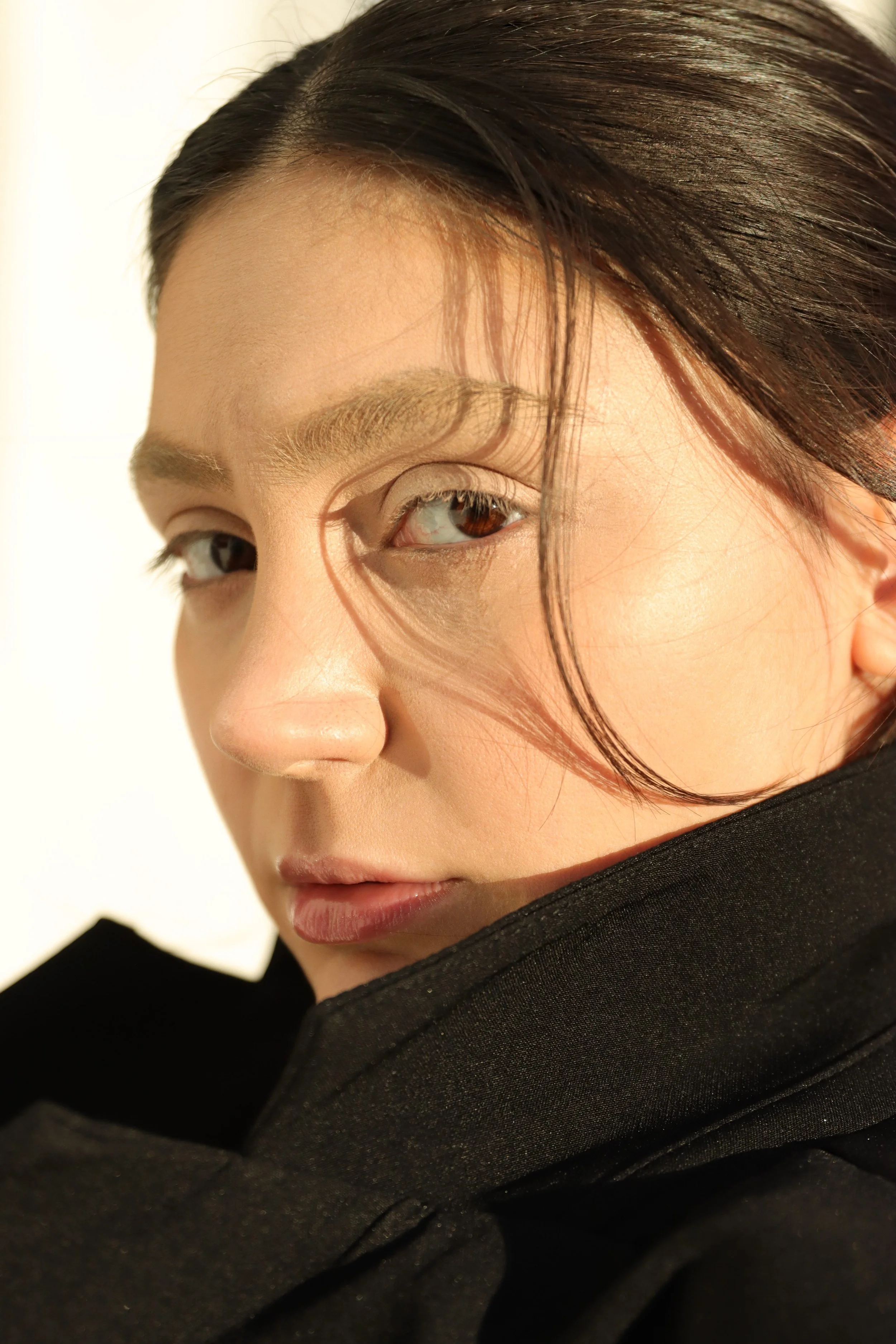 A close-up portrait of a woman with dark hair, partially covering her face, wearing a black jacket or coat, against a plain light background.