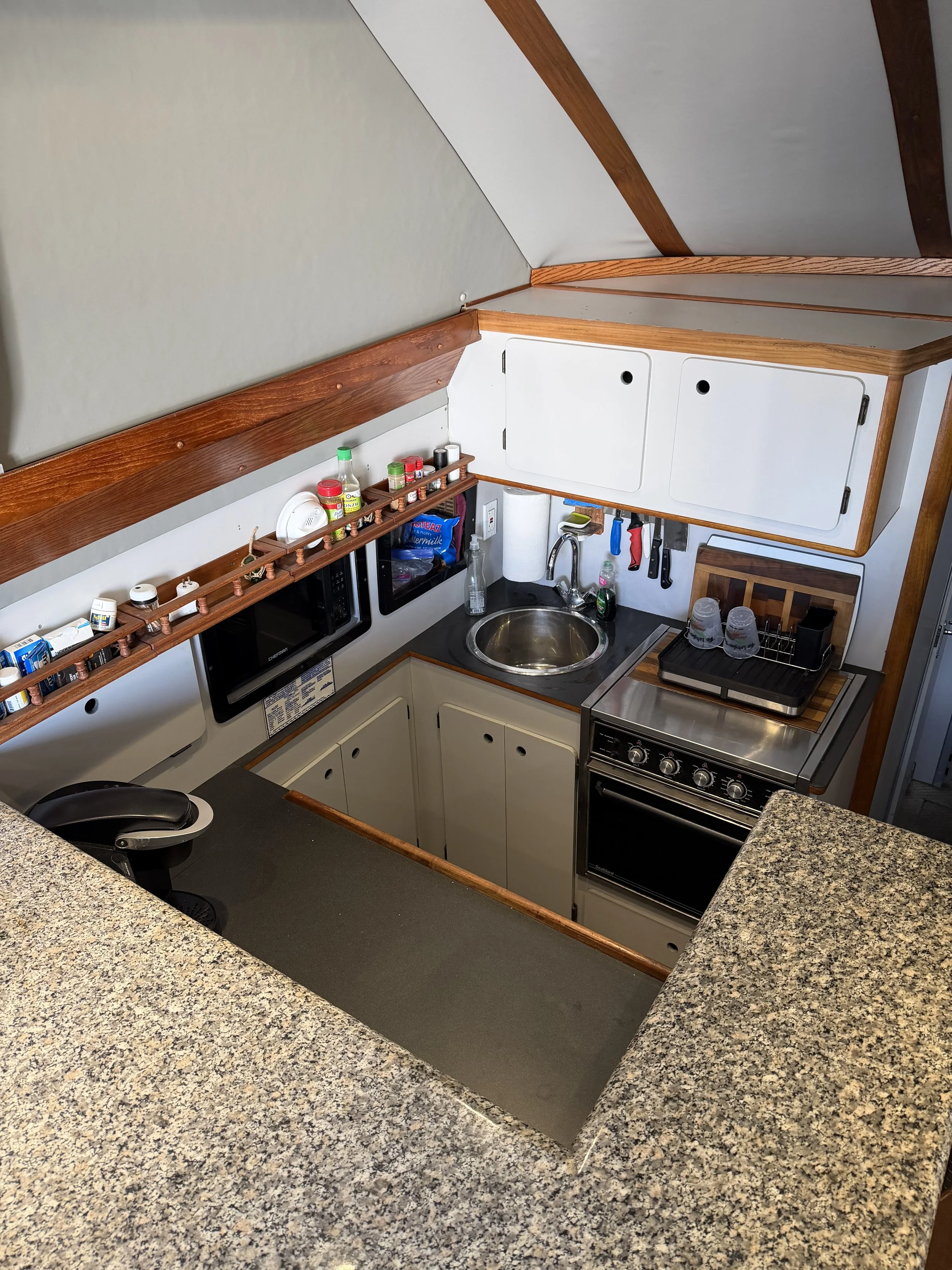 Small kitchen and living area in a boat, featuring a granite countertop, a sink, microwave, cabinets, and a gray sofa.