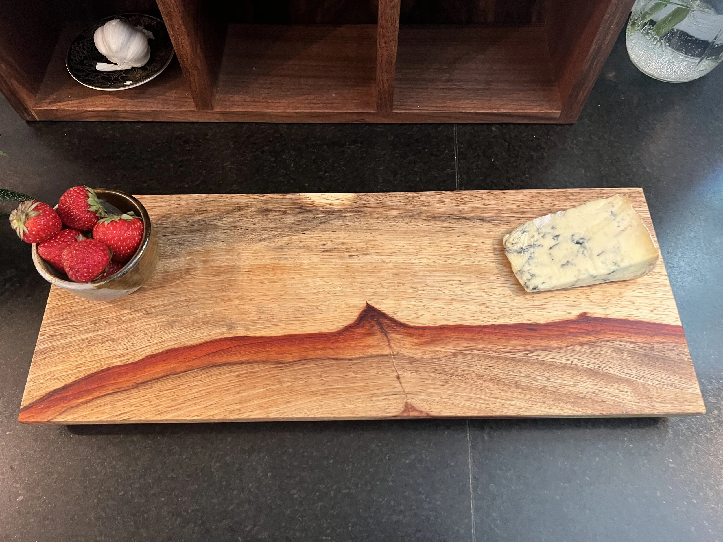 A wooden tray with a bowl of strawberries on the left and a chunk of blue cheese on the right, set on a dark countertop.