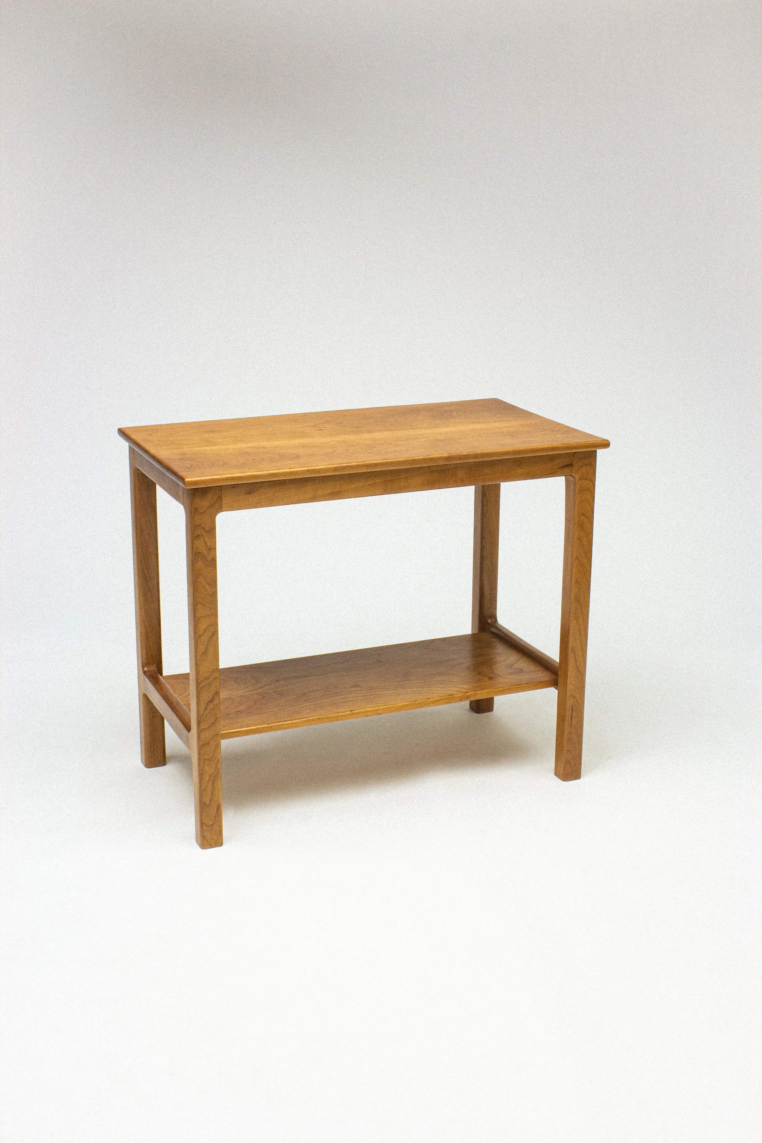 A Cherry wooden mid-century modern table with a lower shelf, set against a plain white background, and handcrafted with wood joinery. 