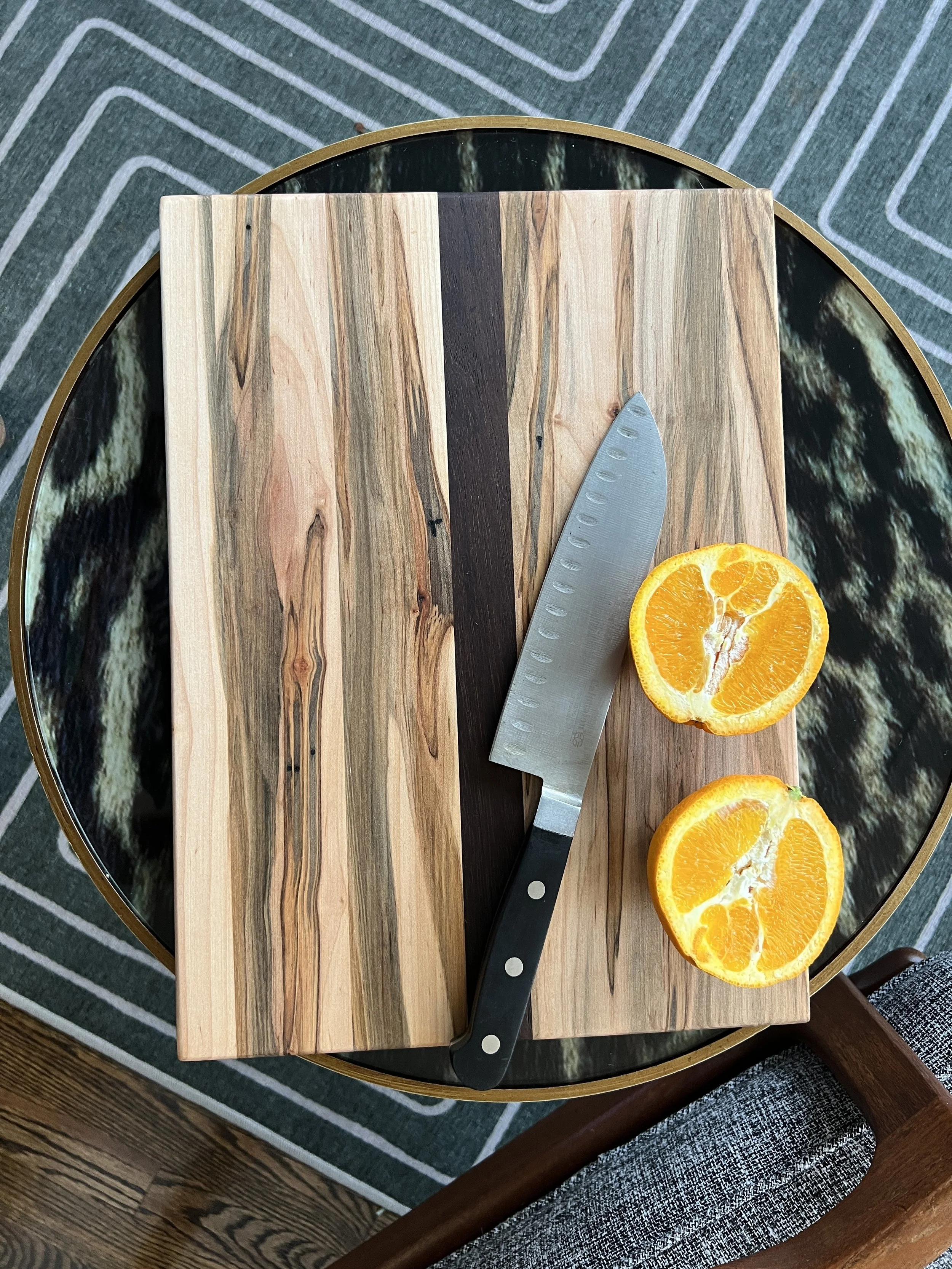Two halved oranges, a kitchen knife, and a wooden cutting board on a round glass table with a patterned green rug below.