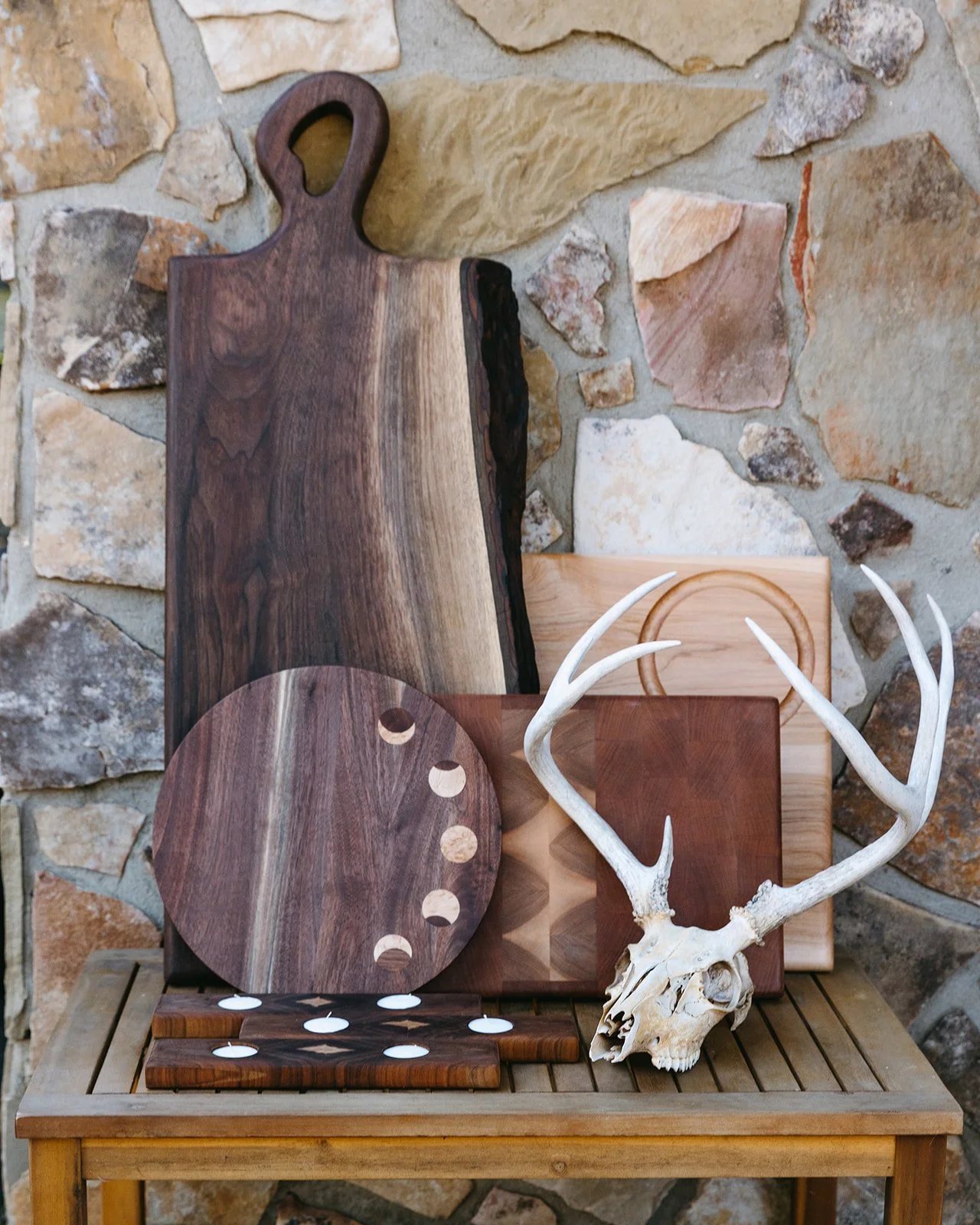 Various wooden cutting boards and decor pieces arranged on a wooden table against a stone wall, including a skull with antlers.