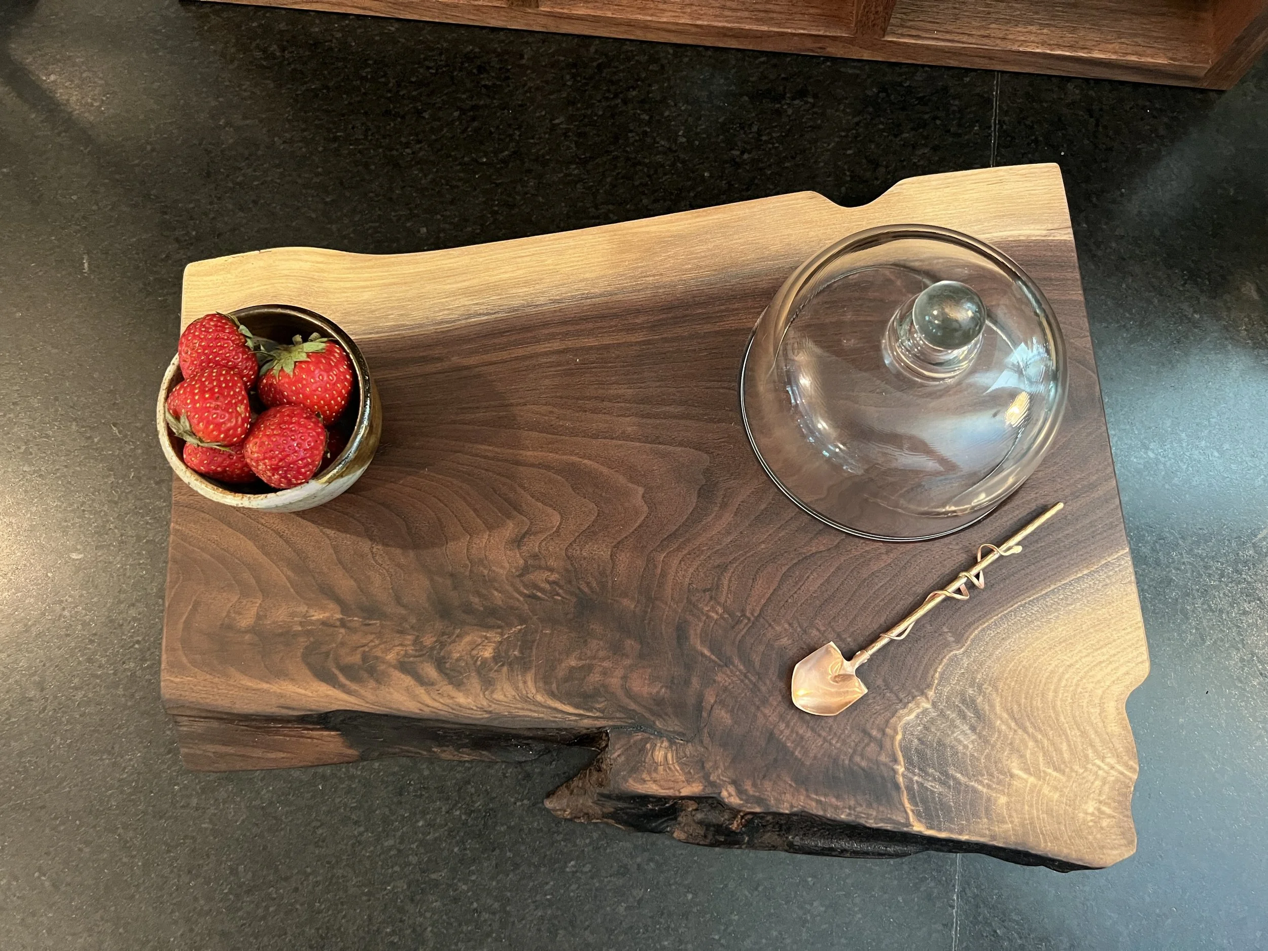 Top-down view of a wooden table with a bowl of strawberries, a clear glass cover, and a gold-colored cocktail stirrer.