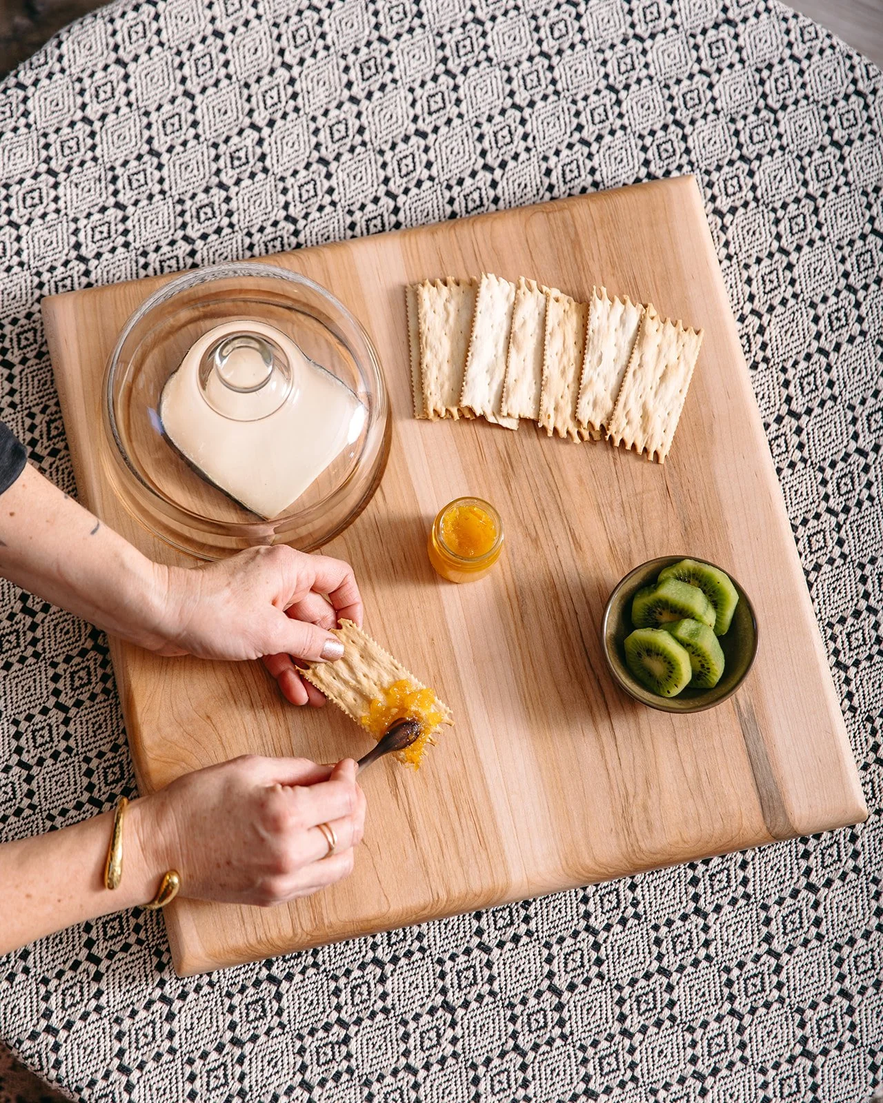 Person spreading orange marmalade on a piece of cracker on a wooden cutting board, surrounded by sliced crackers, a bowl of sliced kiwi, a small jar of orange marmalade, and a glass milk bottle with a cardboard carton inside, with a black and white p