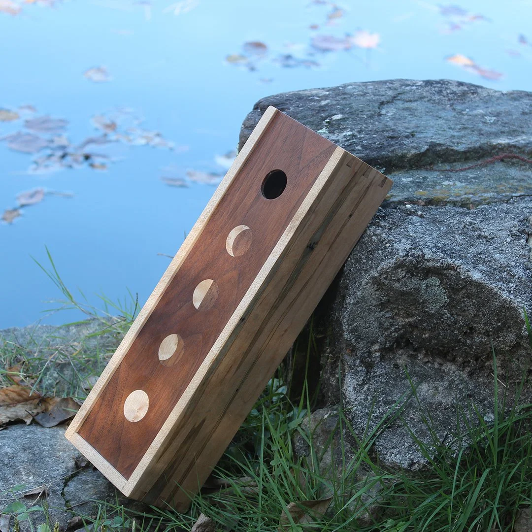 Wooden box made by Toryn Black, woodworker, box is made of walnut, ambosia maple with moon phase inlay vertical on the top. Box is laying against a rock in front of still water 