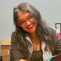 Illustrator and author Nidhi Chanani, with long dark hair wearing red glasses and a dark blazer, smiling while signing one of her books.