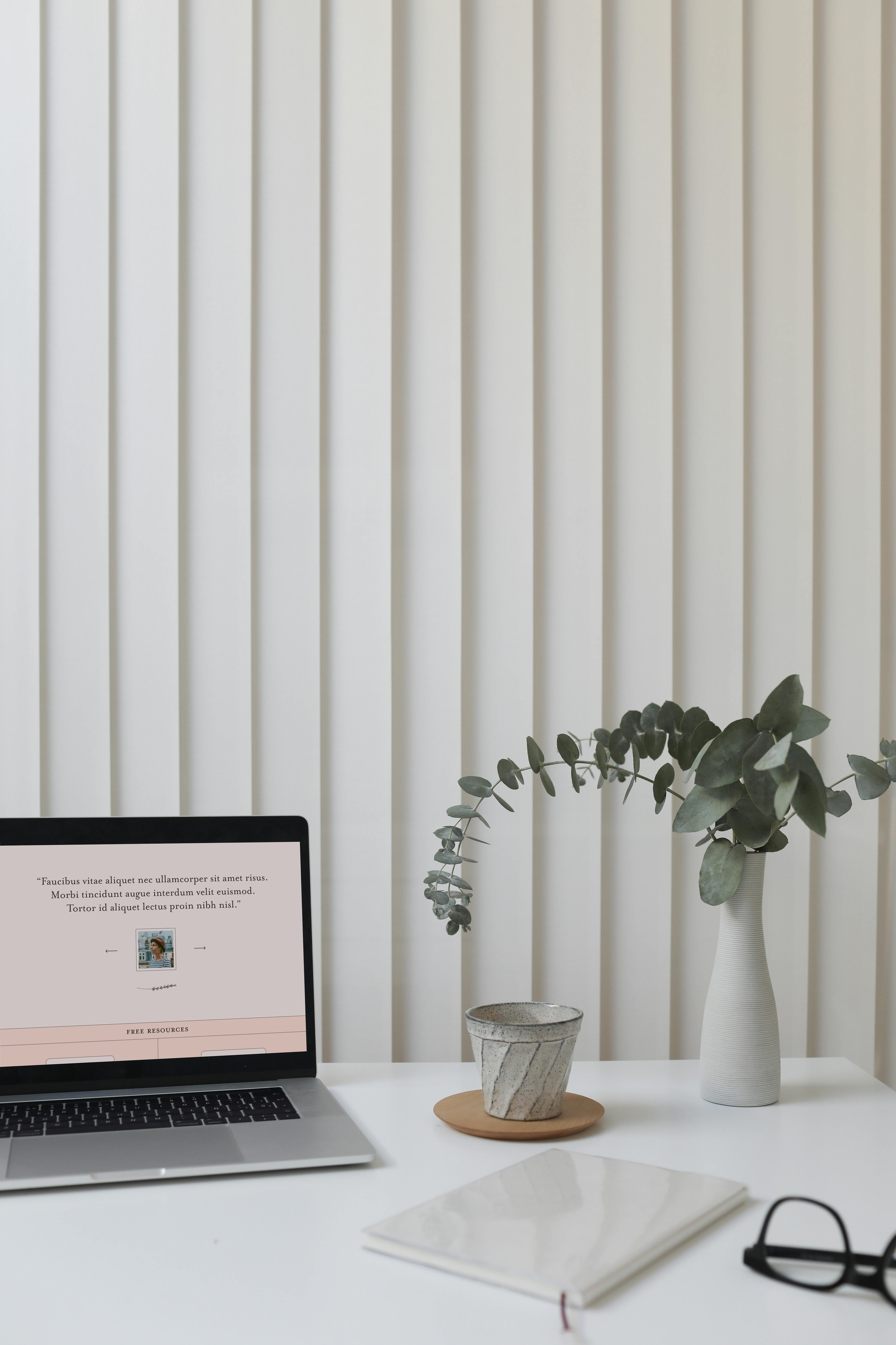 A white desk with an open laptop showing a webpage, a white vase with eucalyptus leaves, a small concrete pot on a round wood tray, a closed notebook, and a pair of glasses, set against a backdrop of white vertical blinds.