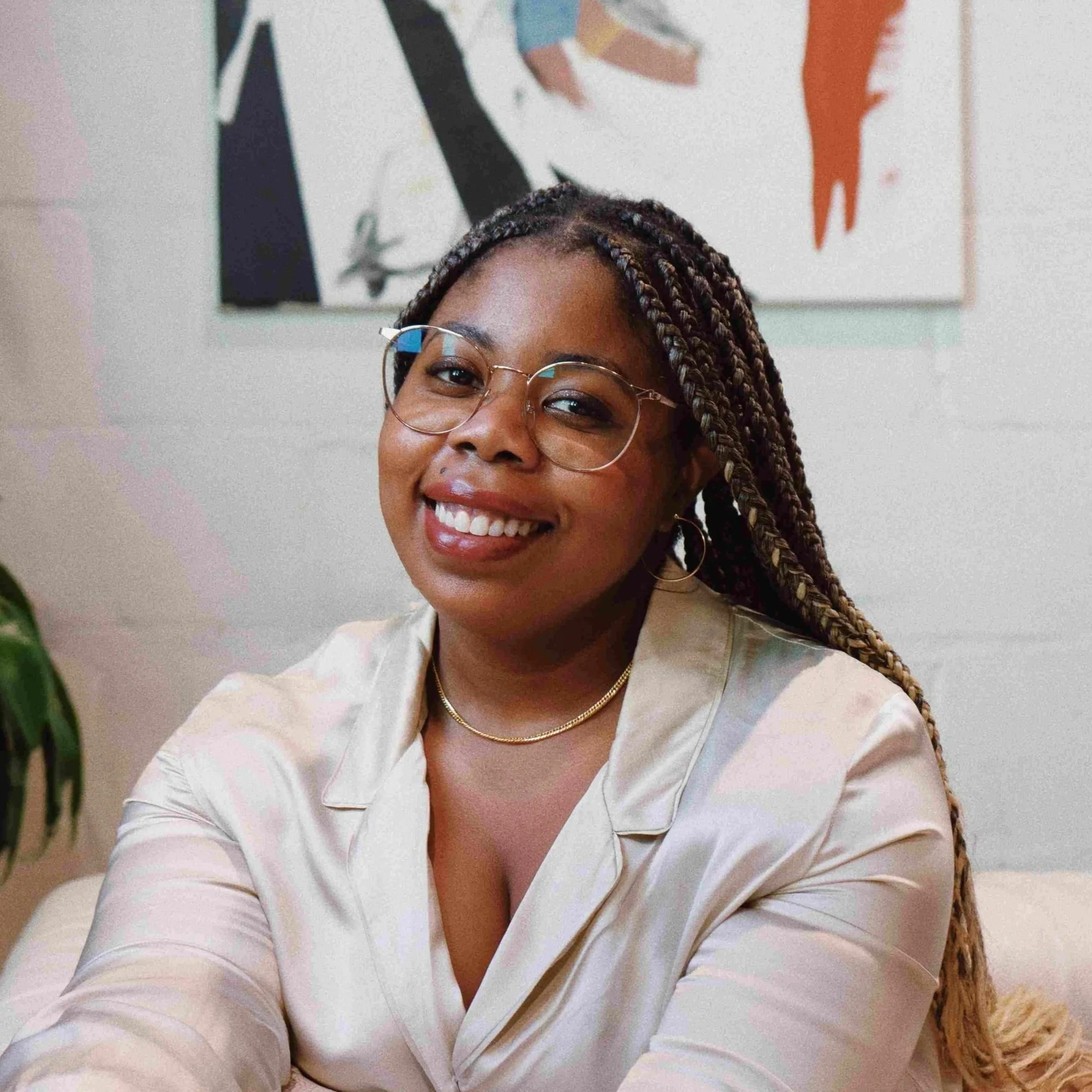 Close-up of Nneka Myers, illustrator and creative director, smiling woman with braided hair, wearing glasses and gold jewelry, in front of an abstract wall art with orange, black, and multicolored shapes.