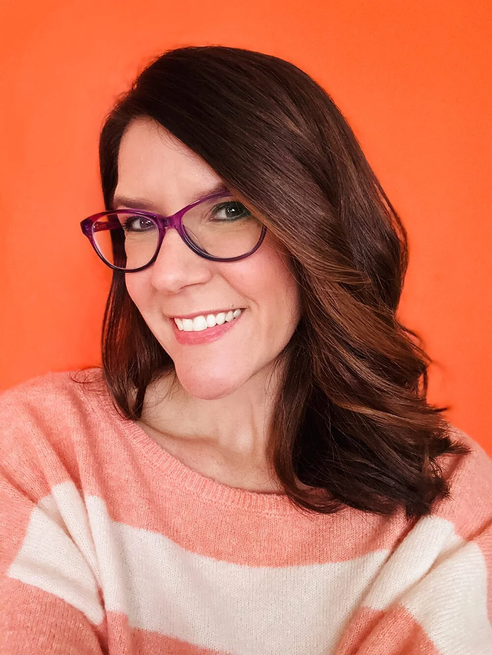 Renee Kurilla, children's book illustrator and author, with wavy brown hair, glasses with purple frames, and a warm smile, wearing a peach and white striped sweater, standing in front of an orange background.