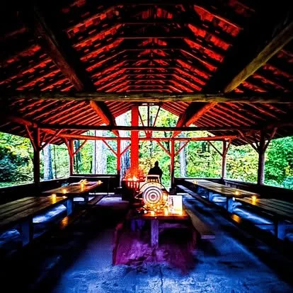 Interior of a wooden pavilion with a domed red roof, long wooden benches along the sides, a table with candles and decorations in the center, and a person sitting at the back against a backdrop of trees and greenery.